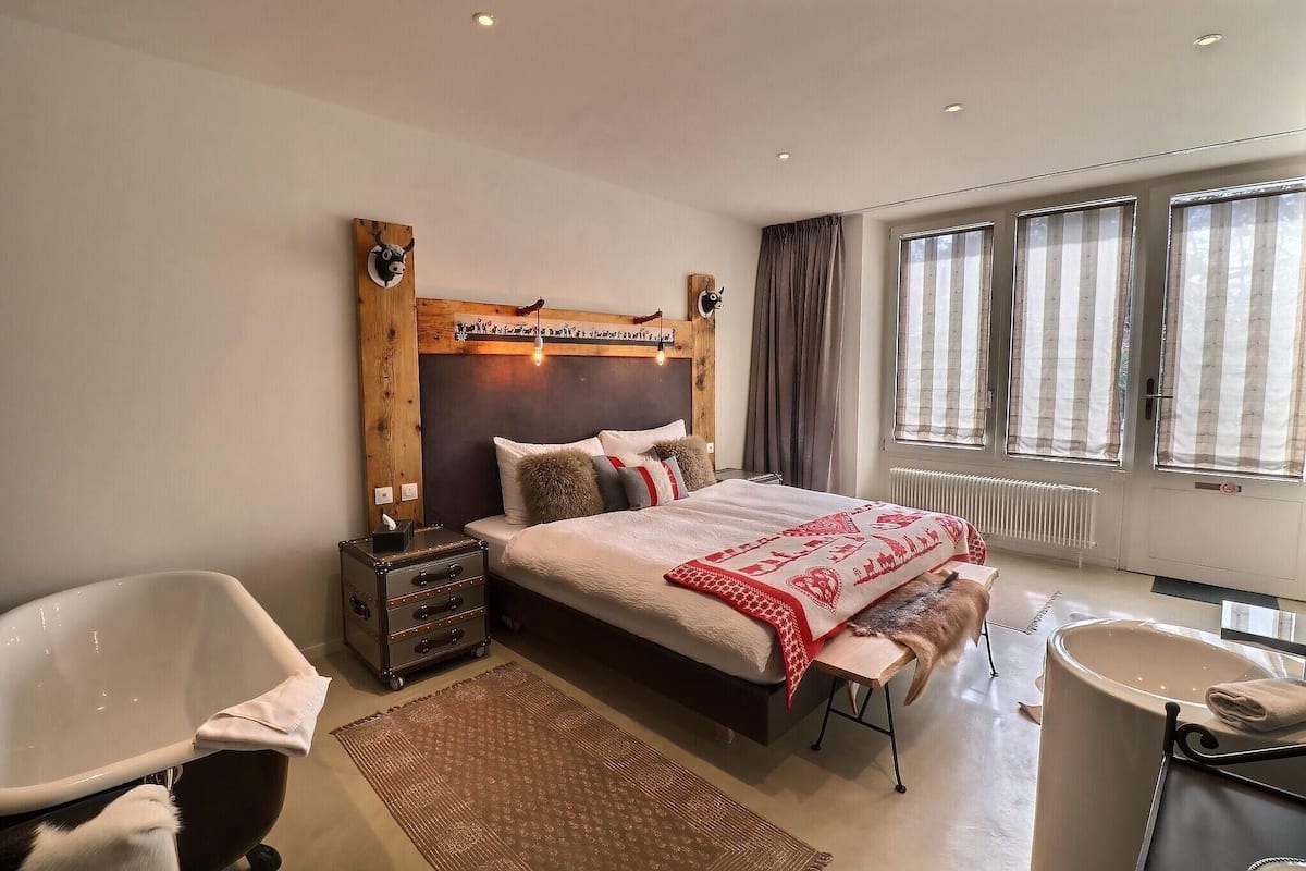 A modern bedroom with a large bed, a wooden headboard, a bathtub, and a large window with curtains.