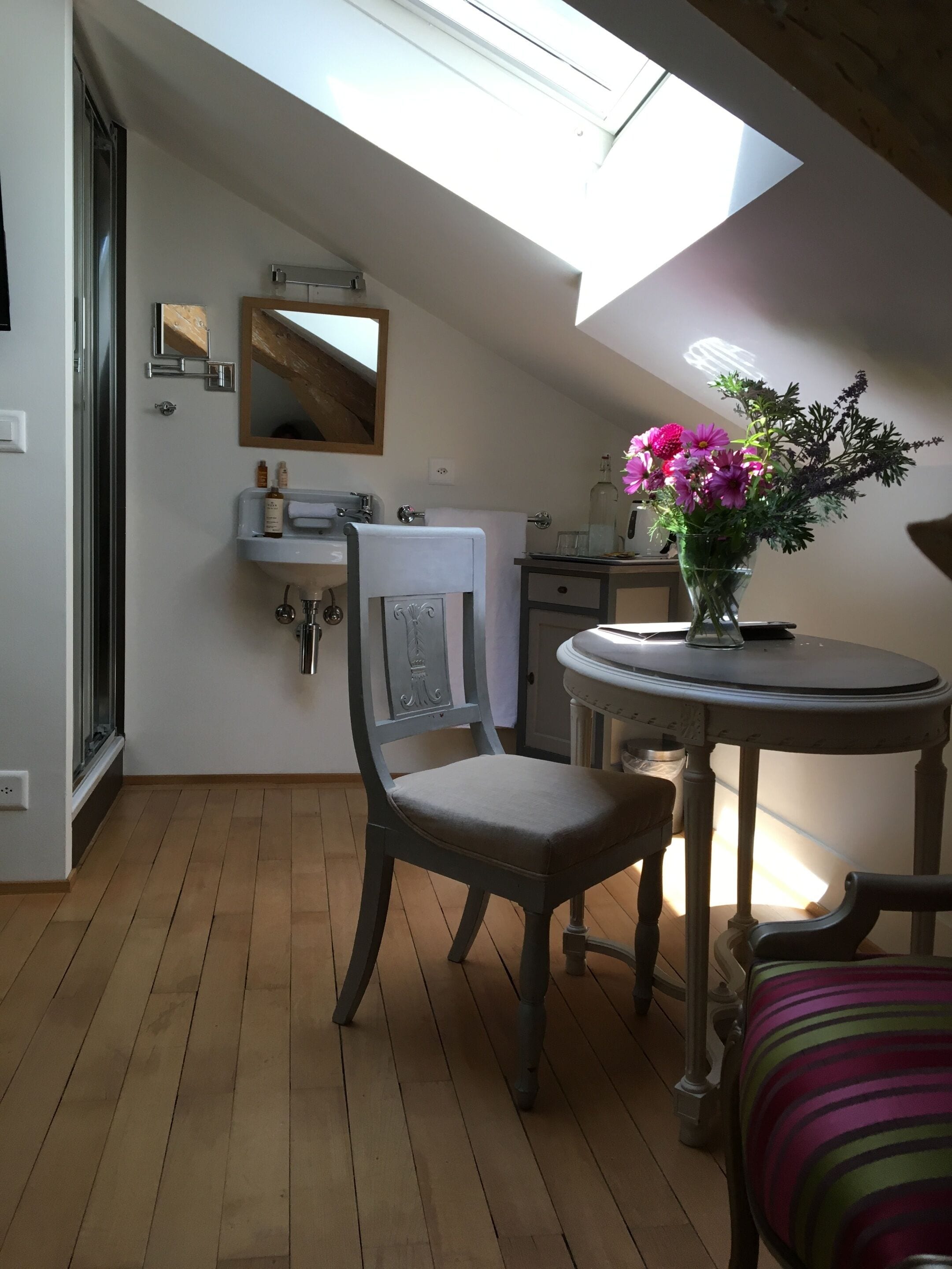 A cozy room with a skylight, a wooden floor, a chair, a table with a vase of flowers, and a sink with toiletries.