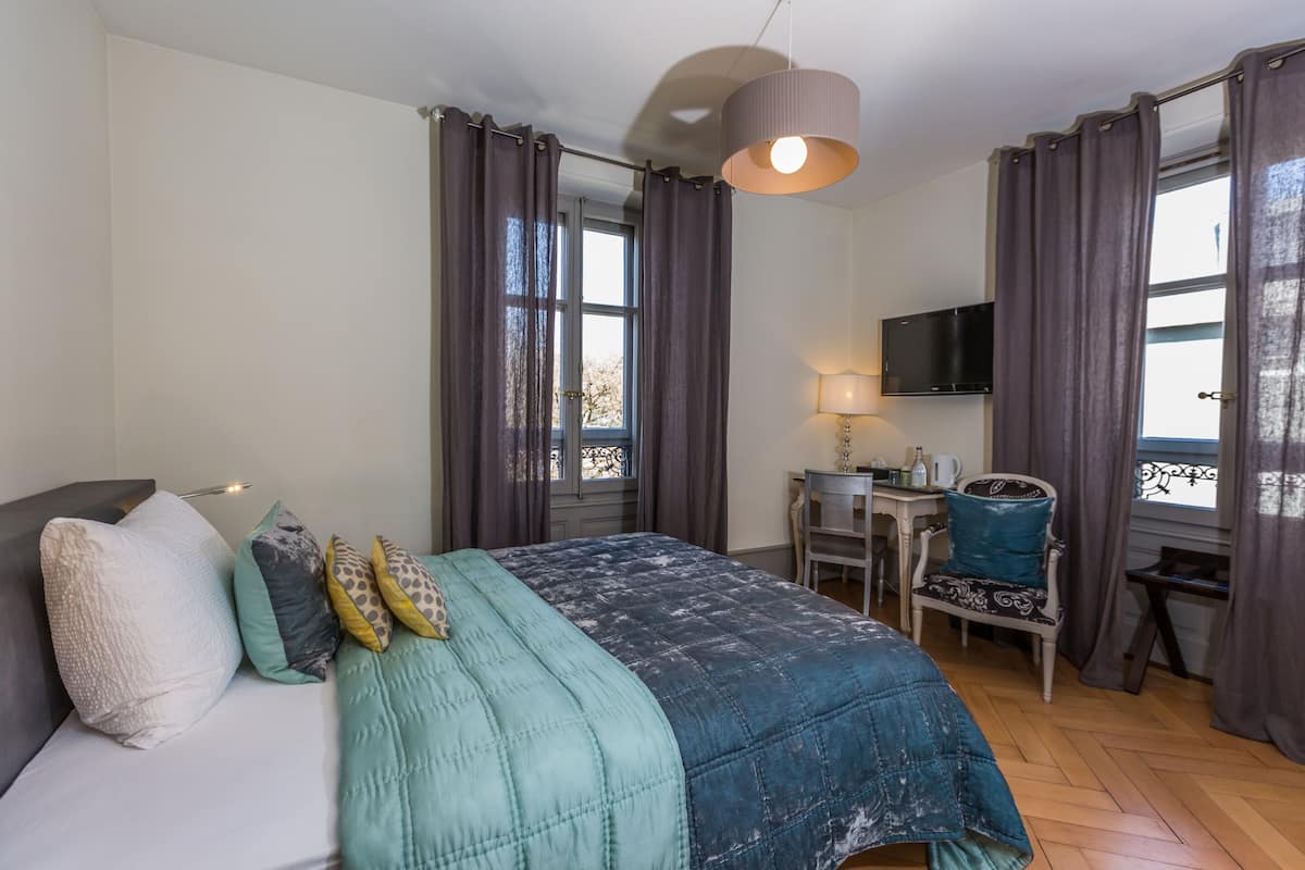 A bedroom with a bed, a desk, a chair, a TV, and a window with curtains.