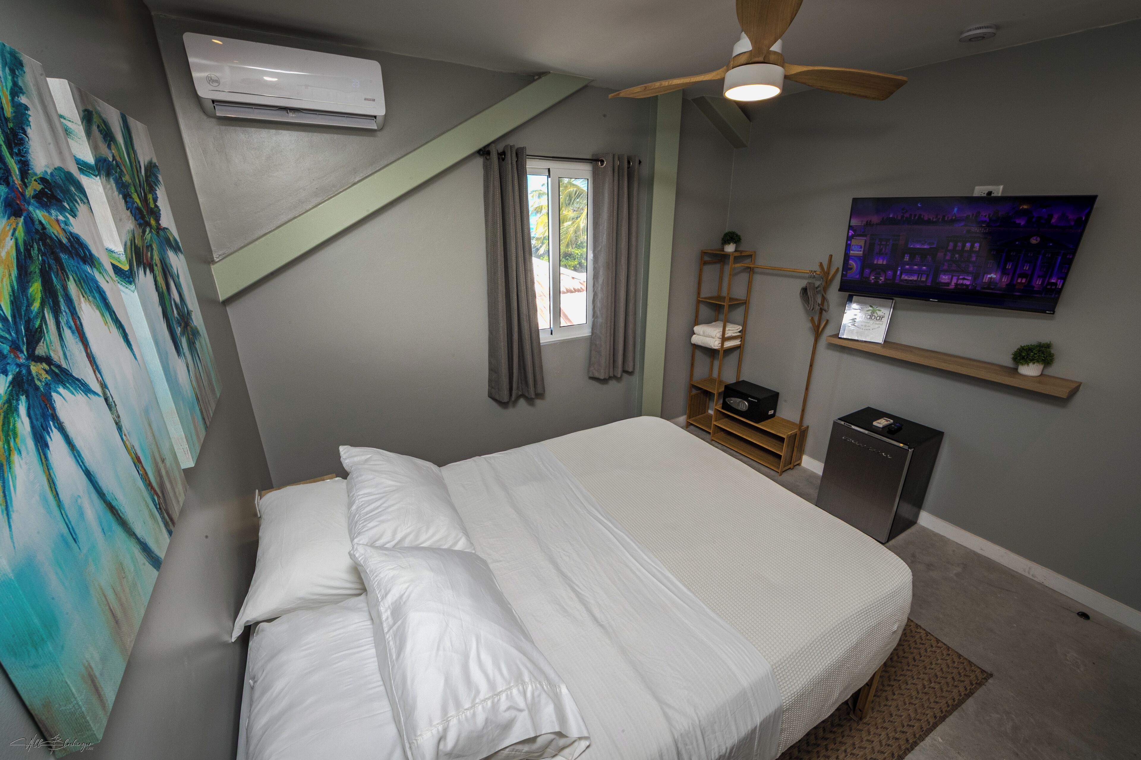 Um quarto com cama, ar condicionado, ventilador de teto, TV, janela com cortinas e um quadro.