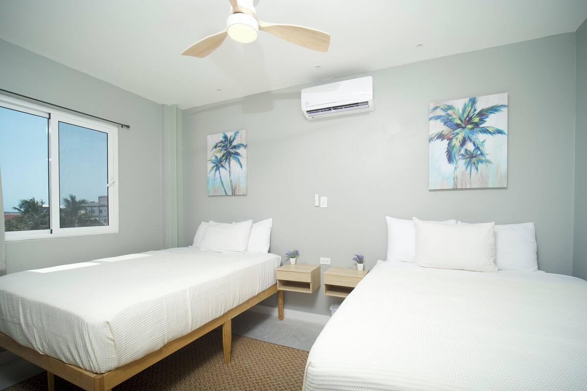 A hotel room with two beds, a ceiling fan, an air conditioning unit, and two wall-mounted paintings of palm trees.