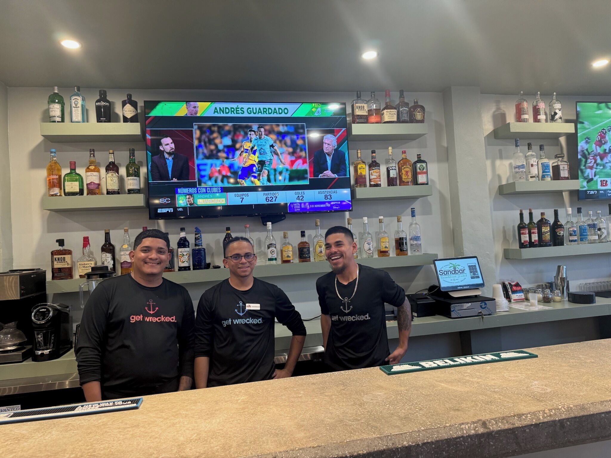 A bar area with multiple shelves of liquor bottles, a television displaying a sports event, and three individuals wearing branded t-shirts.