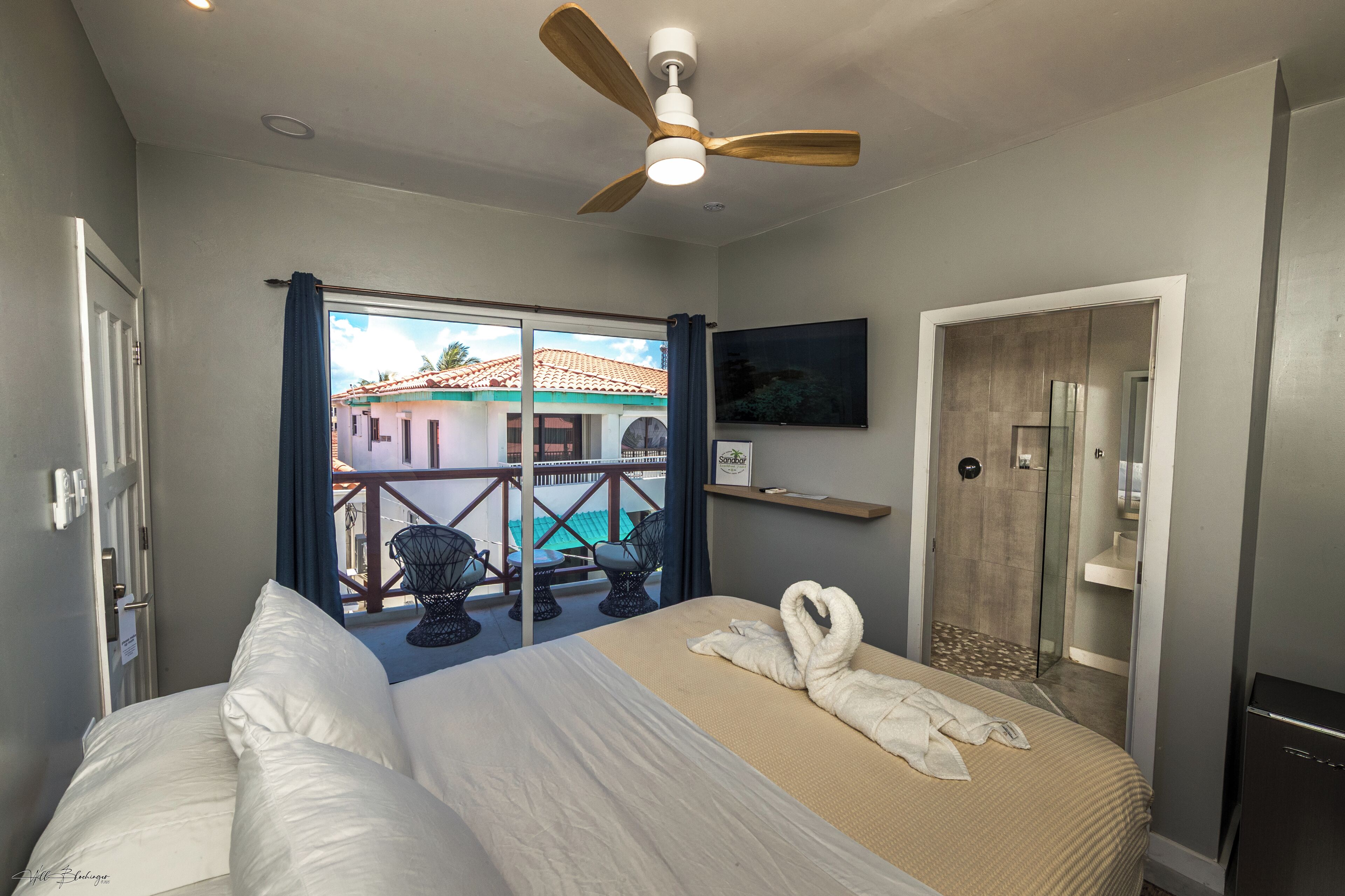 Une chambre d’hôtel avec un lit, un ventilateur de plafond, une douche, un balcon avec des chaises et une vue sur les bâtiments environnants.