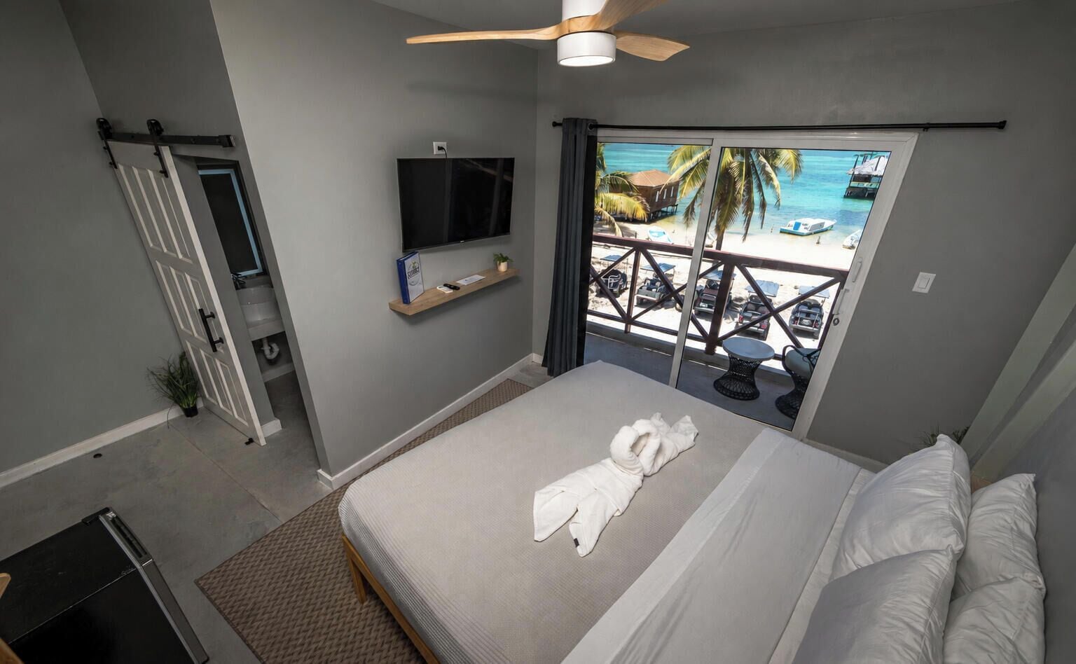 A bedroom with a bed, a TV, a balcony with a view of the beach, and a ceiling fan.