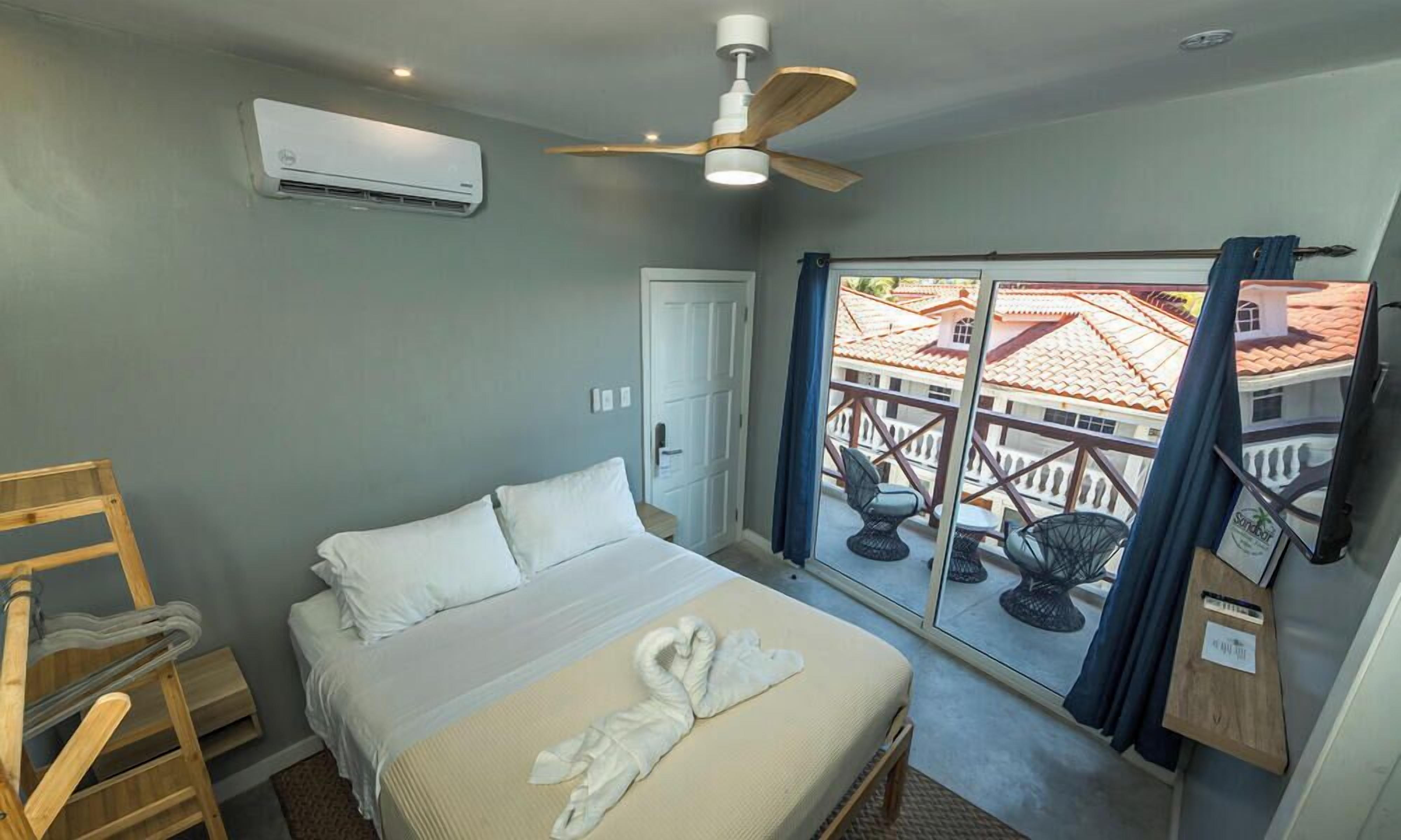 A hotel room with a bed, a ceiling fan, an air conditioning unit, a wooden ladder, a door, and a balcony with outdoor furniture.