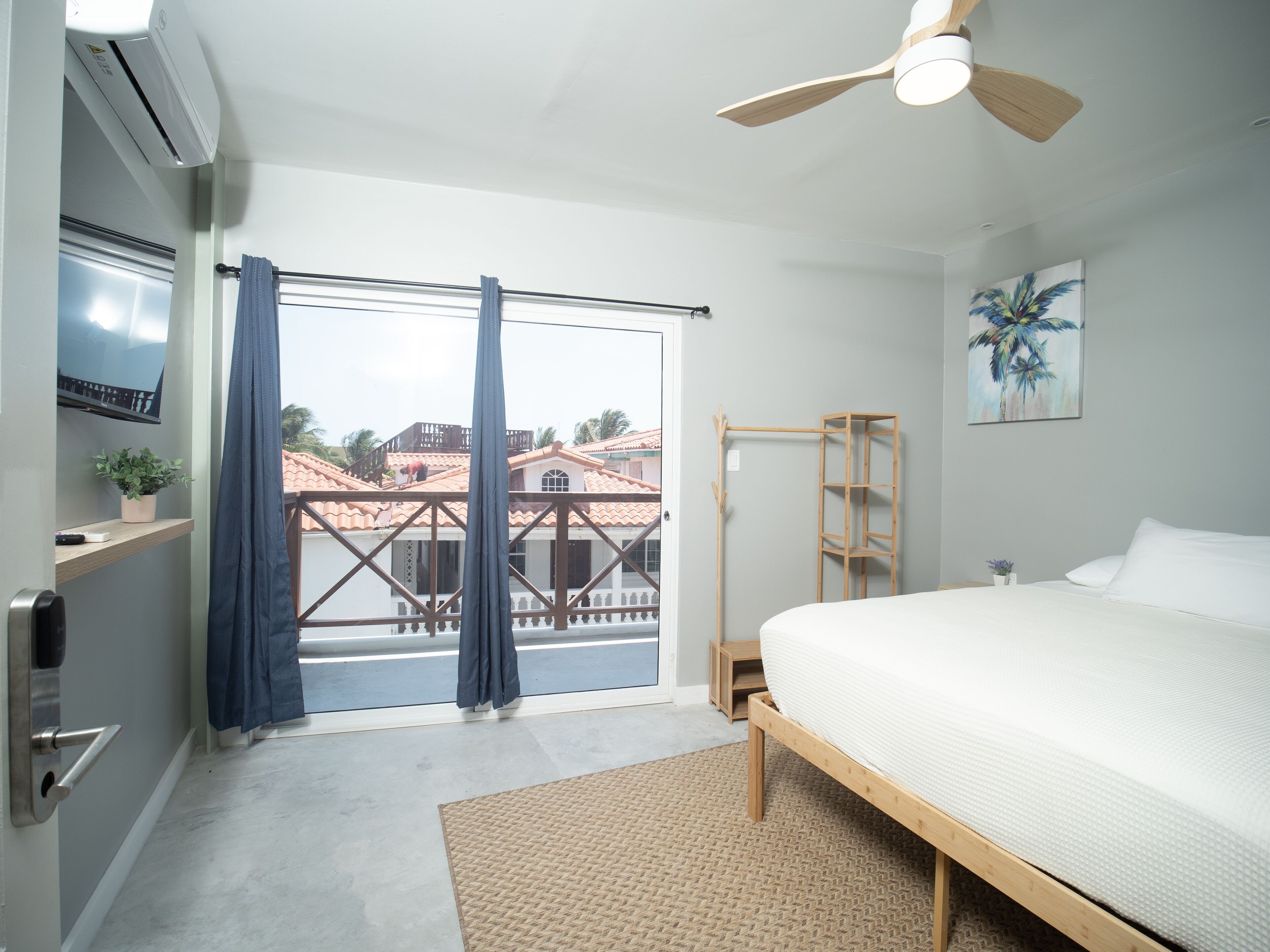 A bedroom with a bed, a ceiling fan, a TV, a window with curtains, and a view of buildings outside.