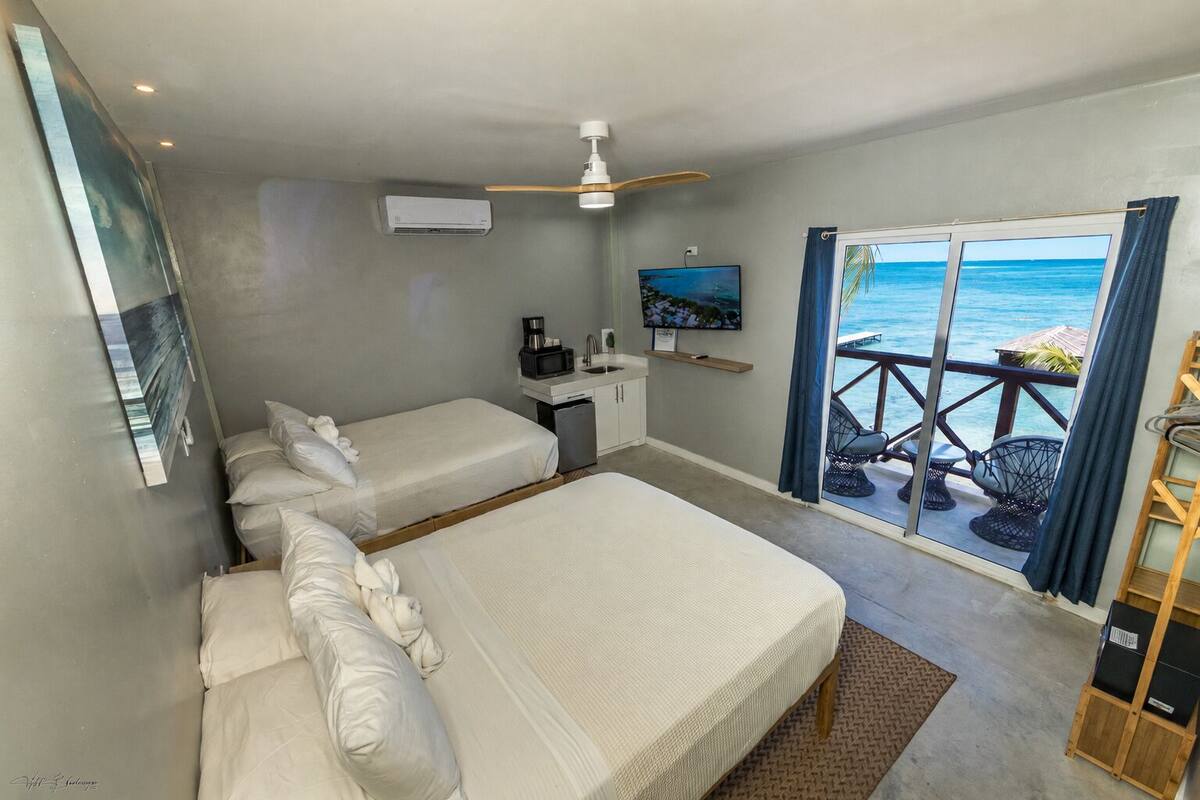 A hotel room with two beds, a ceiling fan, an air conditioning unit, a television, and a balcony with a view of the ocean.