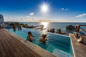A pool with clear water, wooden deck, and a view of the ocean and distant buildings. - Sandbar Beachfront Hotel & Hostel (San Pedro)