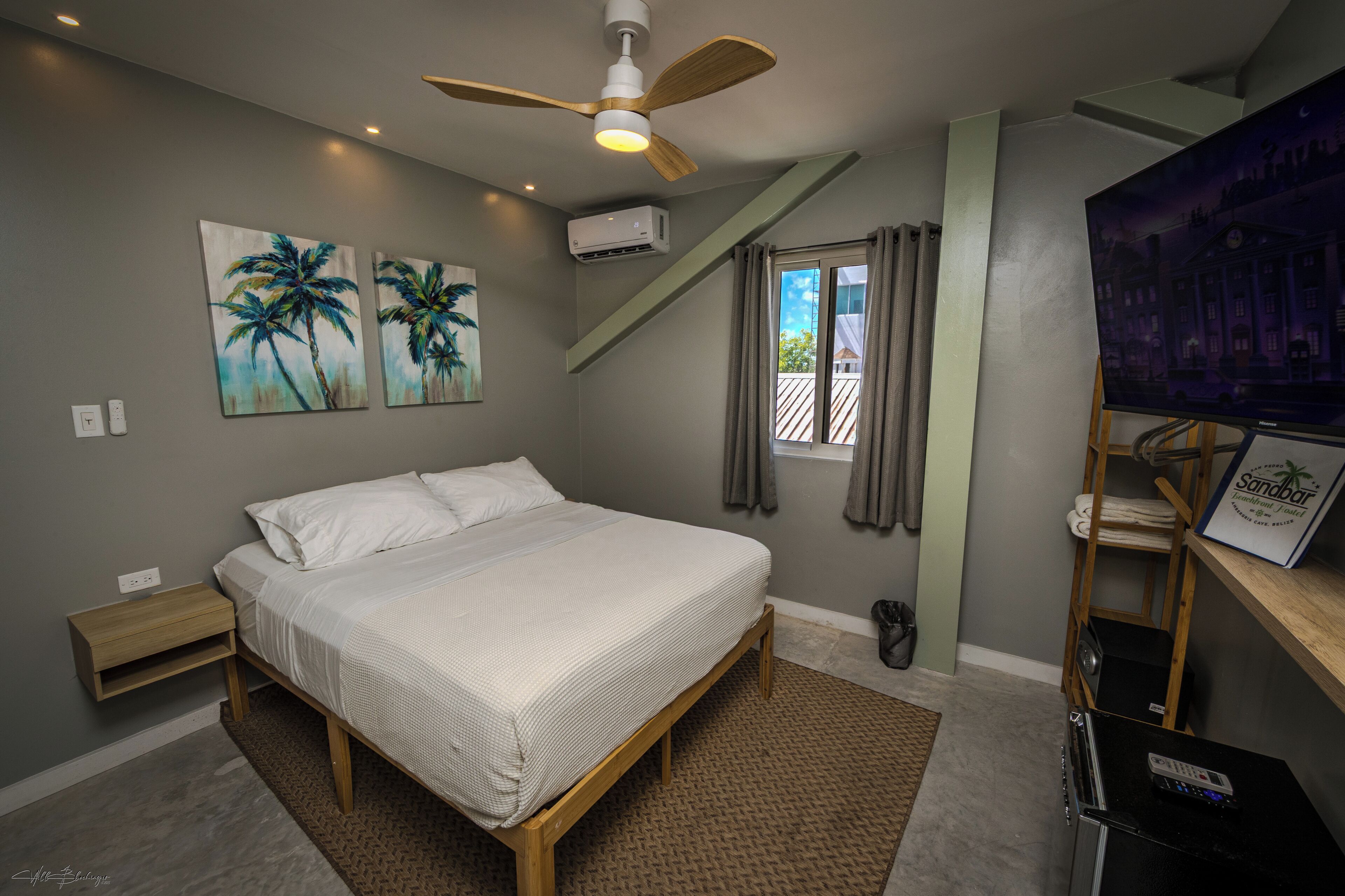 Une chambre à coucher comprenant un lit, un ventilateur de plafond, un appareil de climatisation, une fenêtre avec des rideaux et un téléviseur posé sur un meuble.
