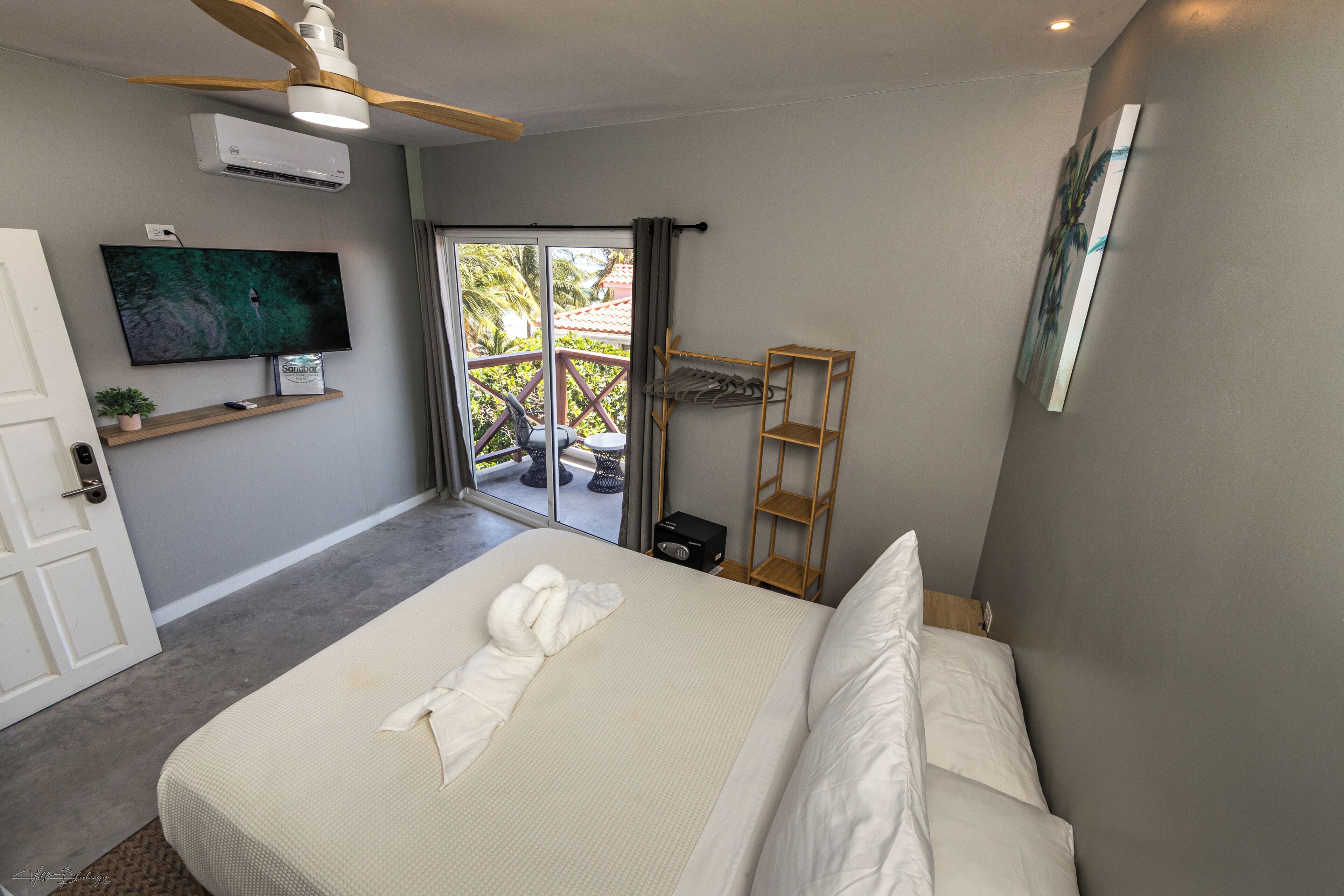 A hotel room with a bed, a TV mounted on the wall, a ceiling fan, and a view of the outdoors through a glass door.