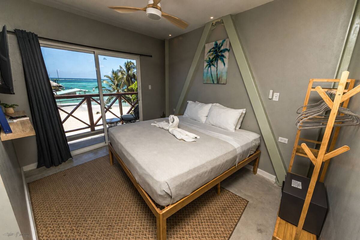 A bedroom with a bed, a view of the ocean, a ceiling fan, and a painting of palm trees.