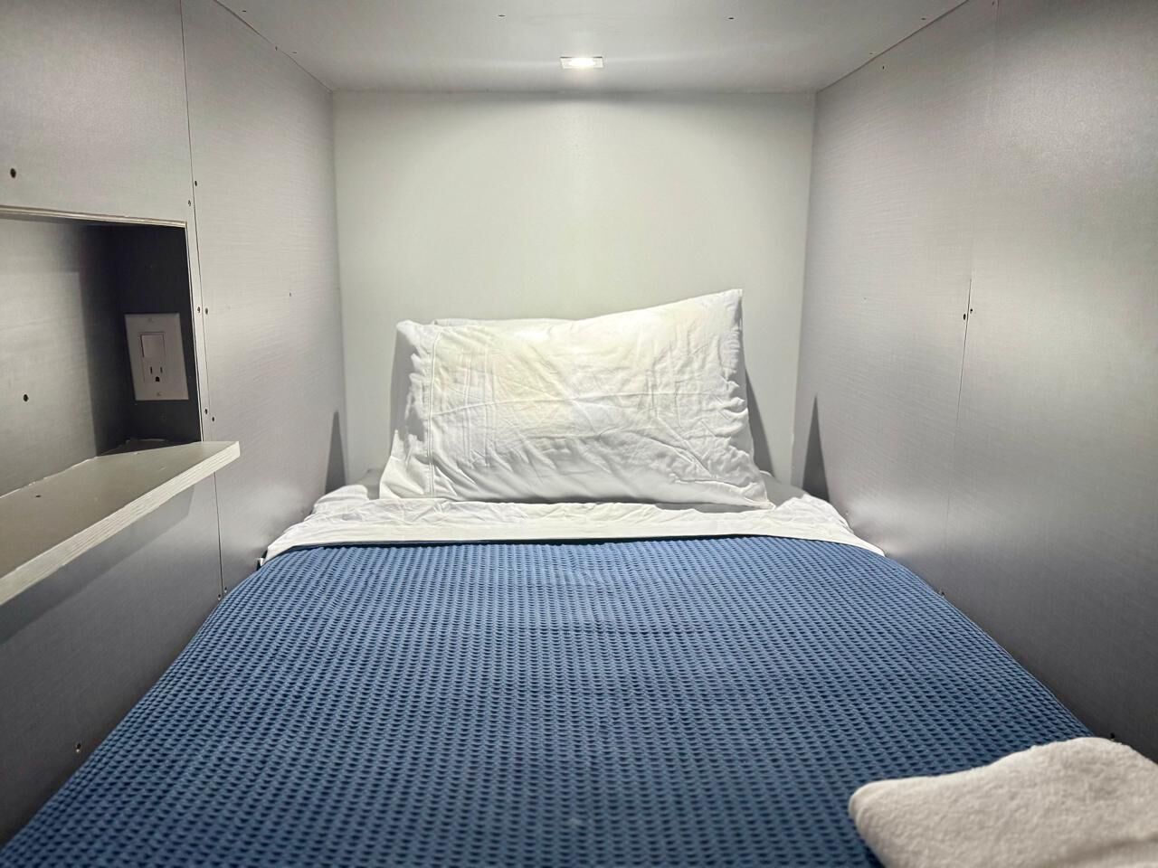 Una habitación pequeña con cama individual, colchón azul, almohada blanca y manta blanca. En la pared izquierda hay una estantería con un enchufe.