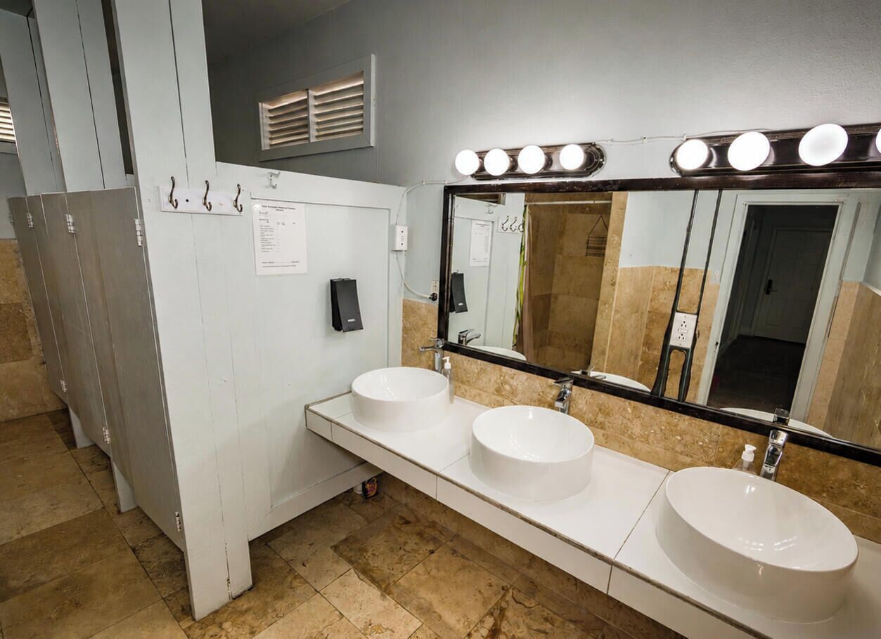 A restroom with three sinks, a mirror with lights, and a ventilation system.