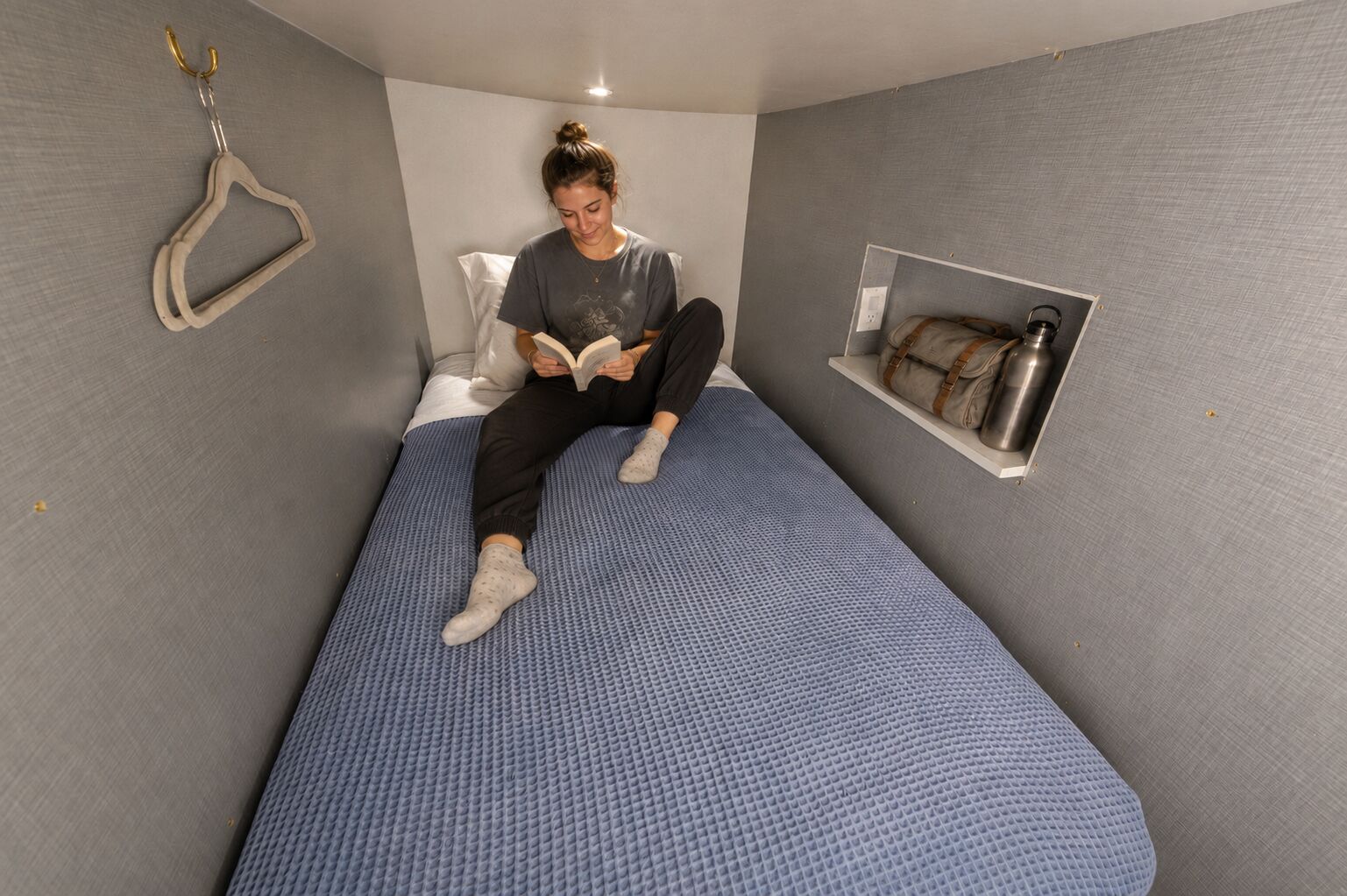 Una habitación pequeña con cama individual, colchón azul, almohada blanca, pared gris, estante con una botella de agua y un gancho en la pared.