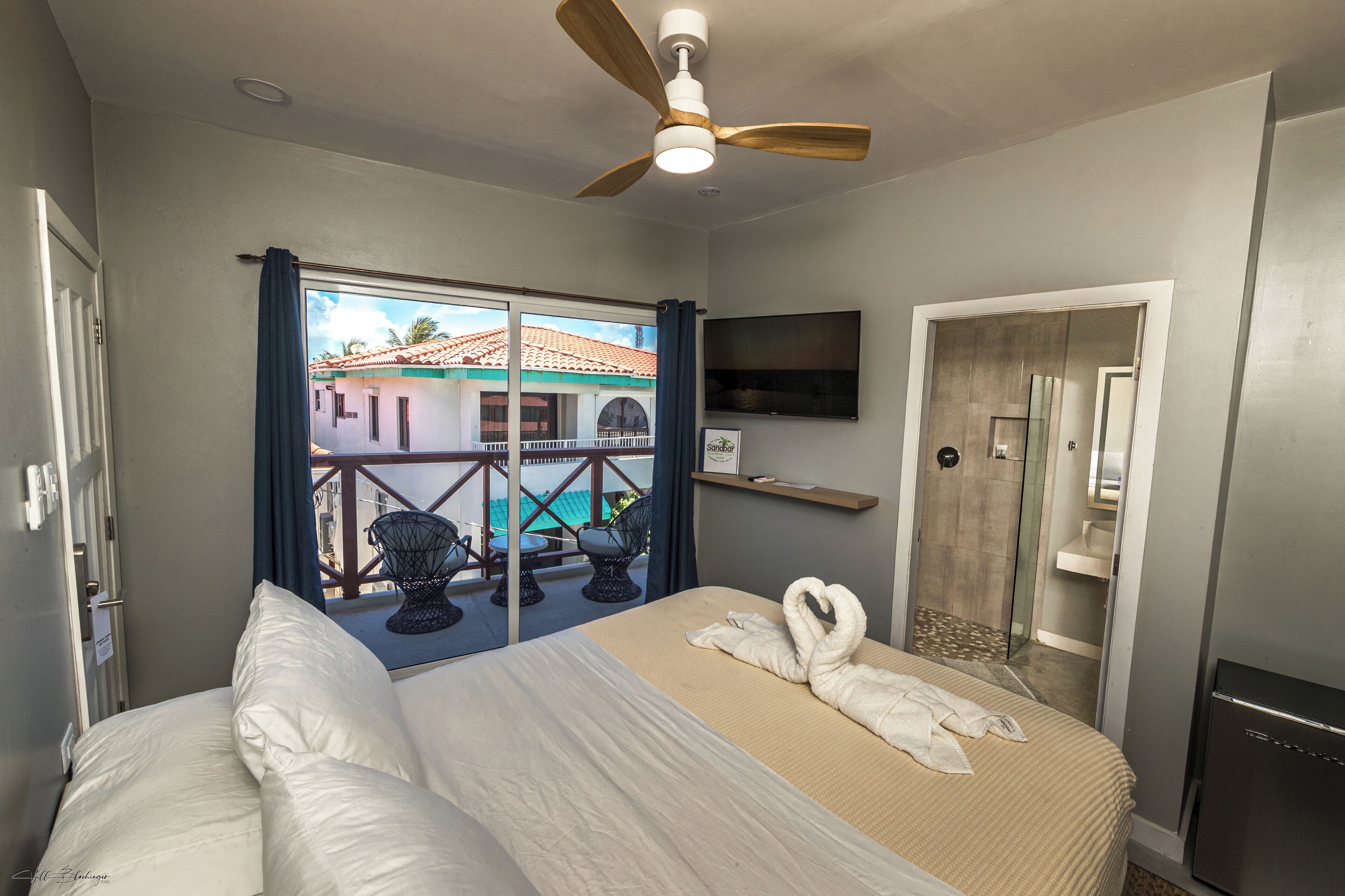 Habitación de hotel con cama, ventilador de techo, televisión, ducha y balcón con vista a edificios.