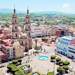 Un paisaje urbano con edificios históricos destacados, incluyendo una catedral con dos torres y una cúpula central. Se observa una glorieta con una fuente y abundante vegetación bien cuidada.