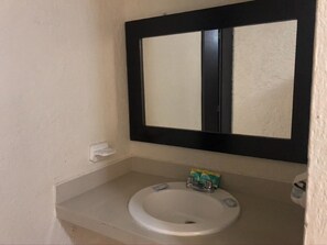 Un baño con un lavamanos blanco, un espejo con marco negro y un estante pequeño con artículos de aseo.