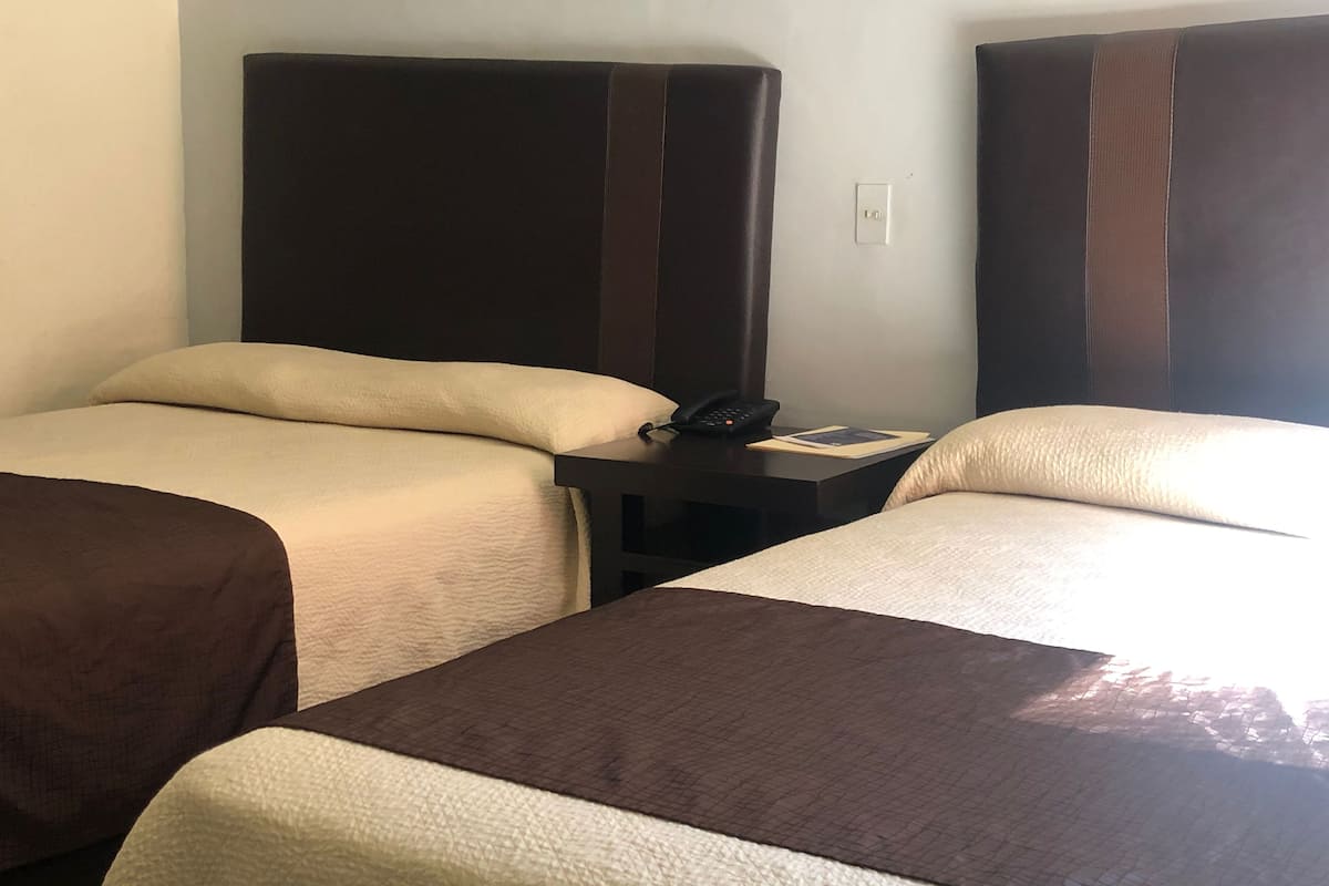 Habitación de hotel con dos camas, cada una con cabecero marrón, una mesita de noche con teléfono e interruptor de luz en la pared.