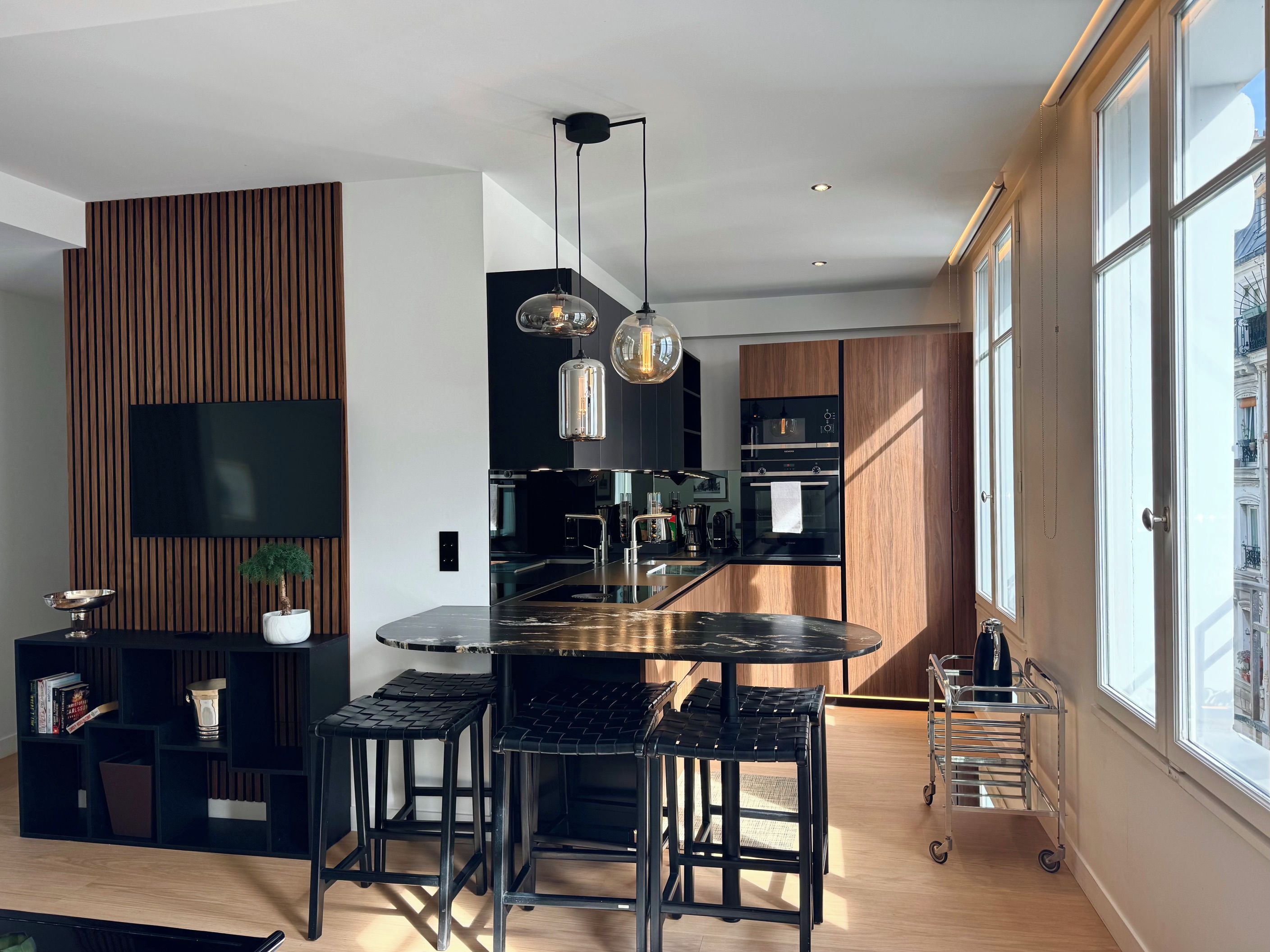 A modern kitchen with a central island, black bar stools, a flat-screen TV mounted on a wooden panel, and a view of the city through large windows.