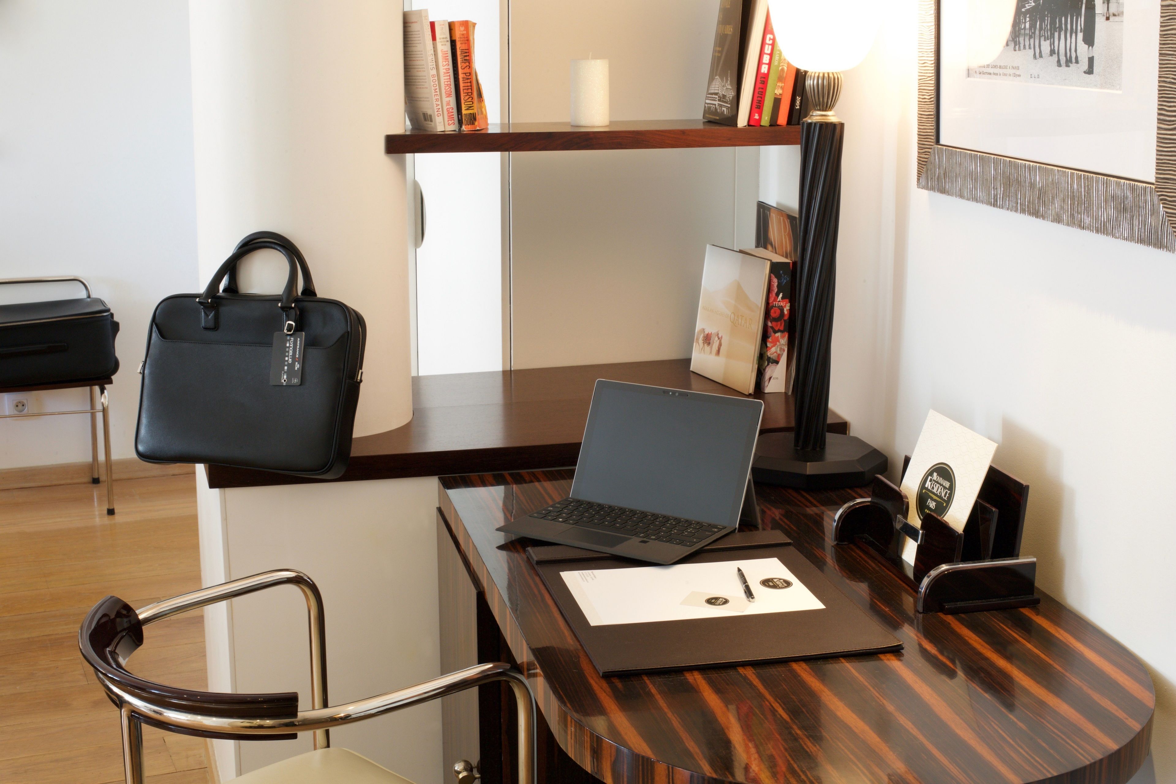 Un bureau en bois avec un ordinateur portable, un sac à main noir, un tableau encadré et une bibliothèque contenant des livres et une bougie.