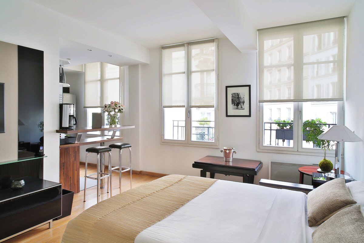 A modern hotel room with a large bed, a kitchenette, a dining area, and large windows.