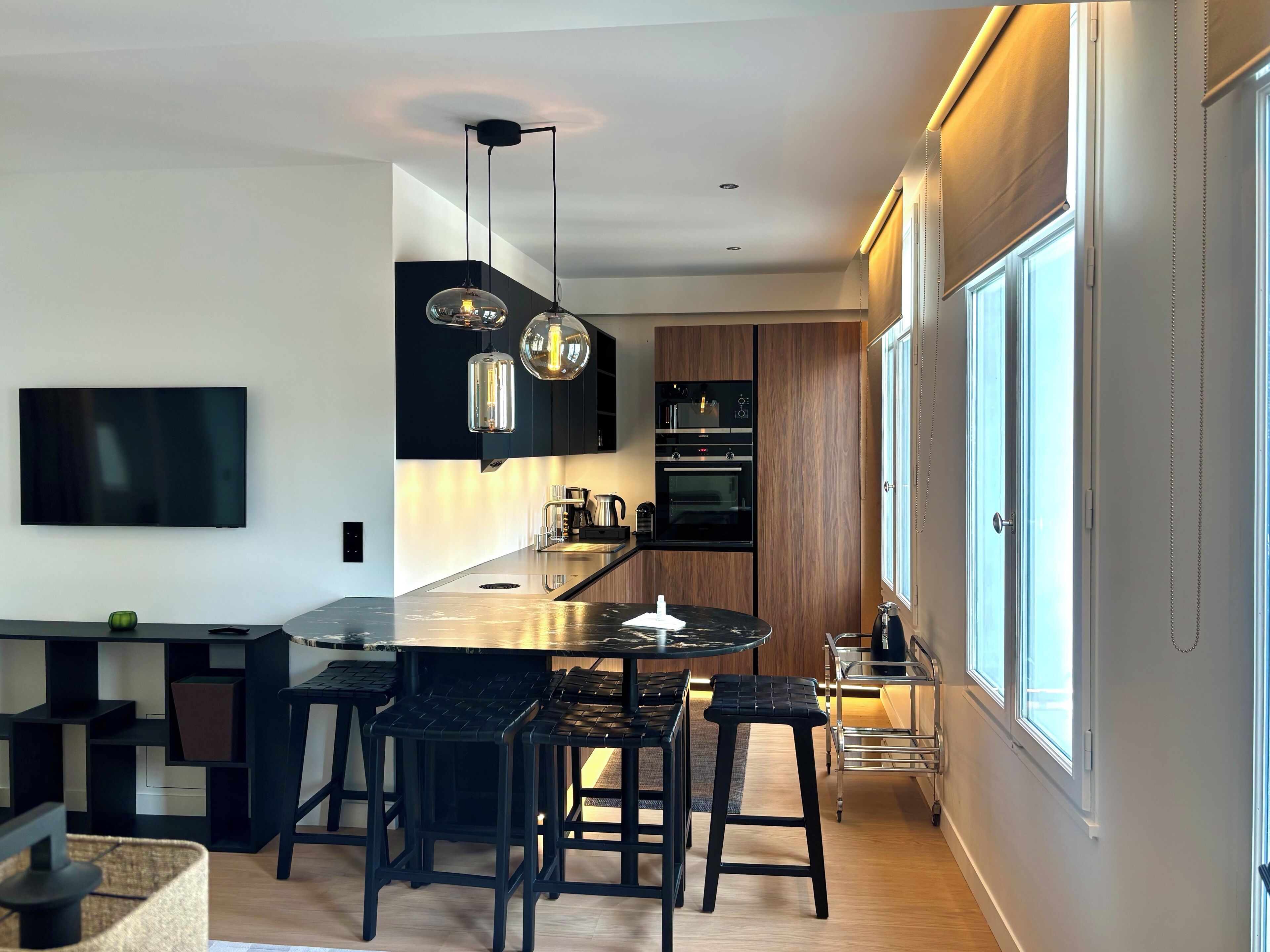 Una cocina moderna con una isla central, taburetes de bar, iluminación colgante y un amplio ventanal.