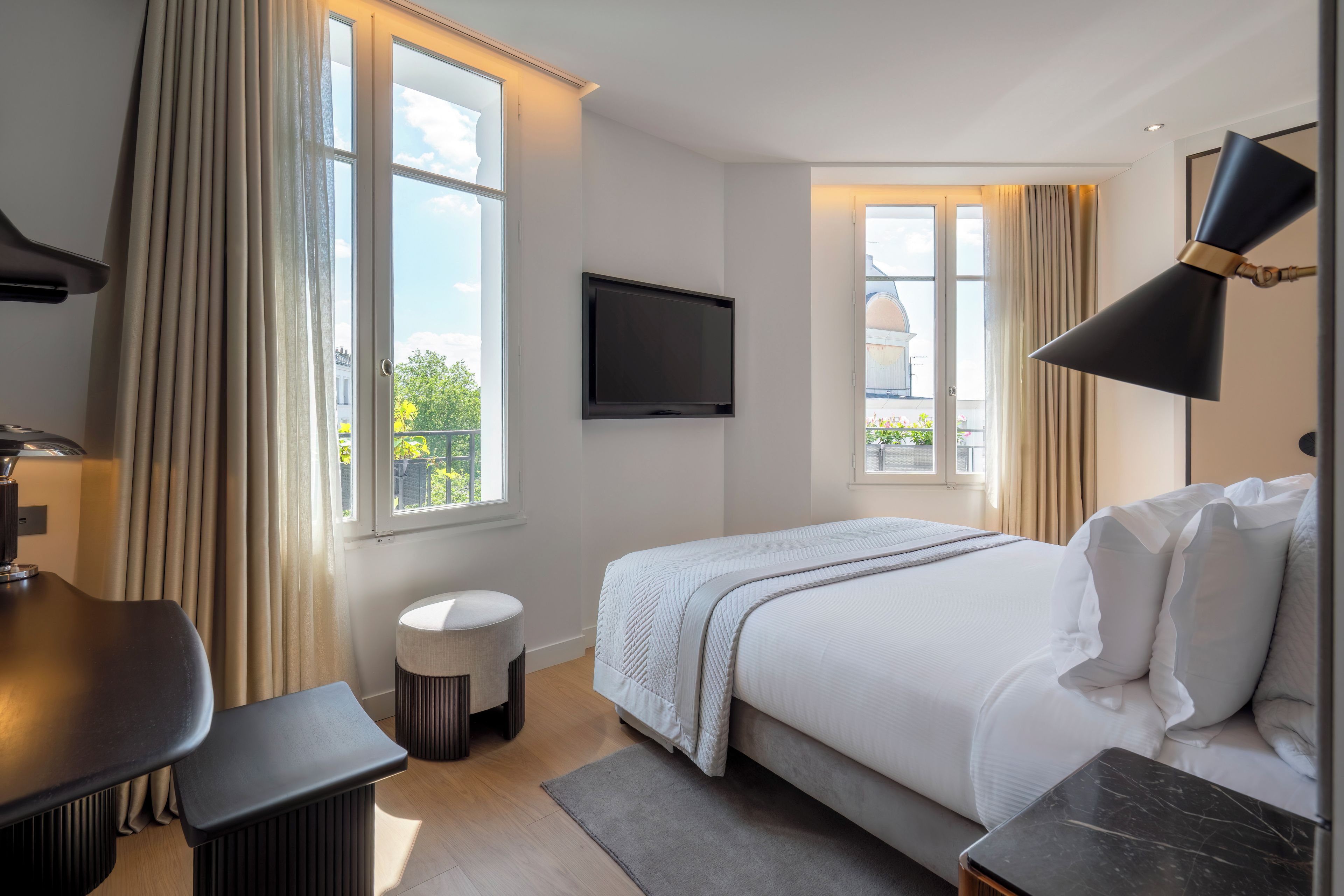 A modern hotel room with a bed, a desk, a TV, and large windows.