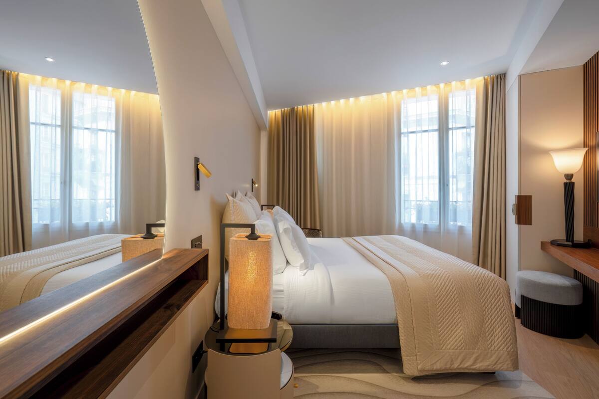 Habitación de hotel con dos camas, una mesita de noche, una lámpara y un ventanal con cortinas.