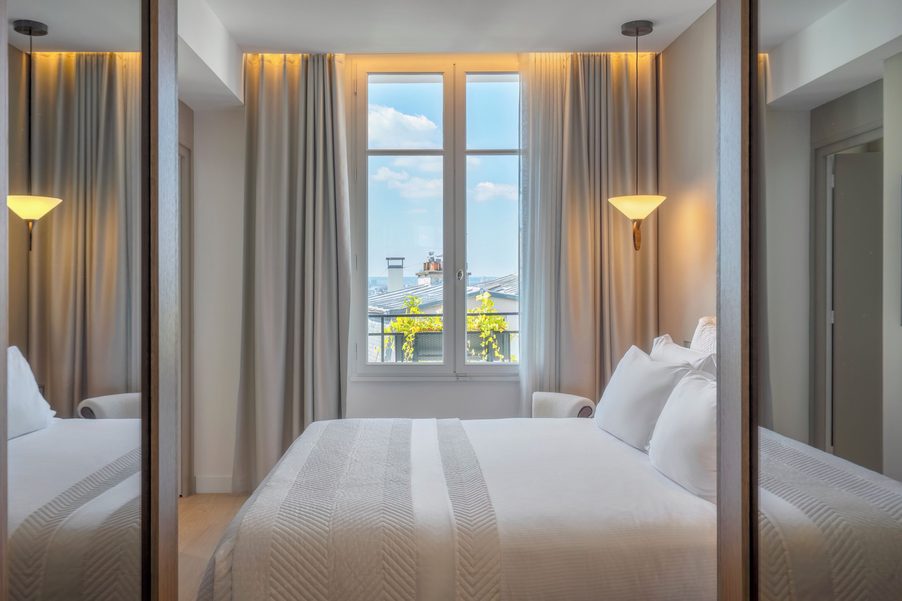 Ett hotellrum med en stor säng, två sängbord, ett fönster med gardiner och utsikt över byggnader och träd.