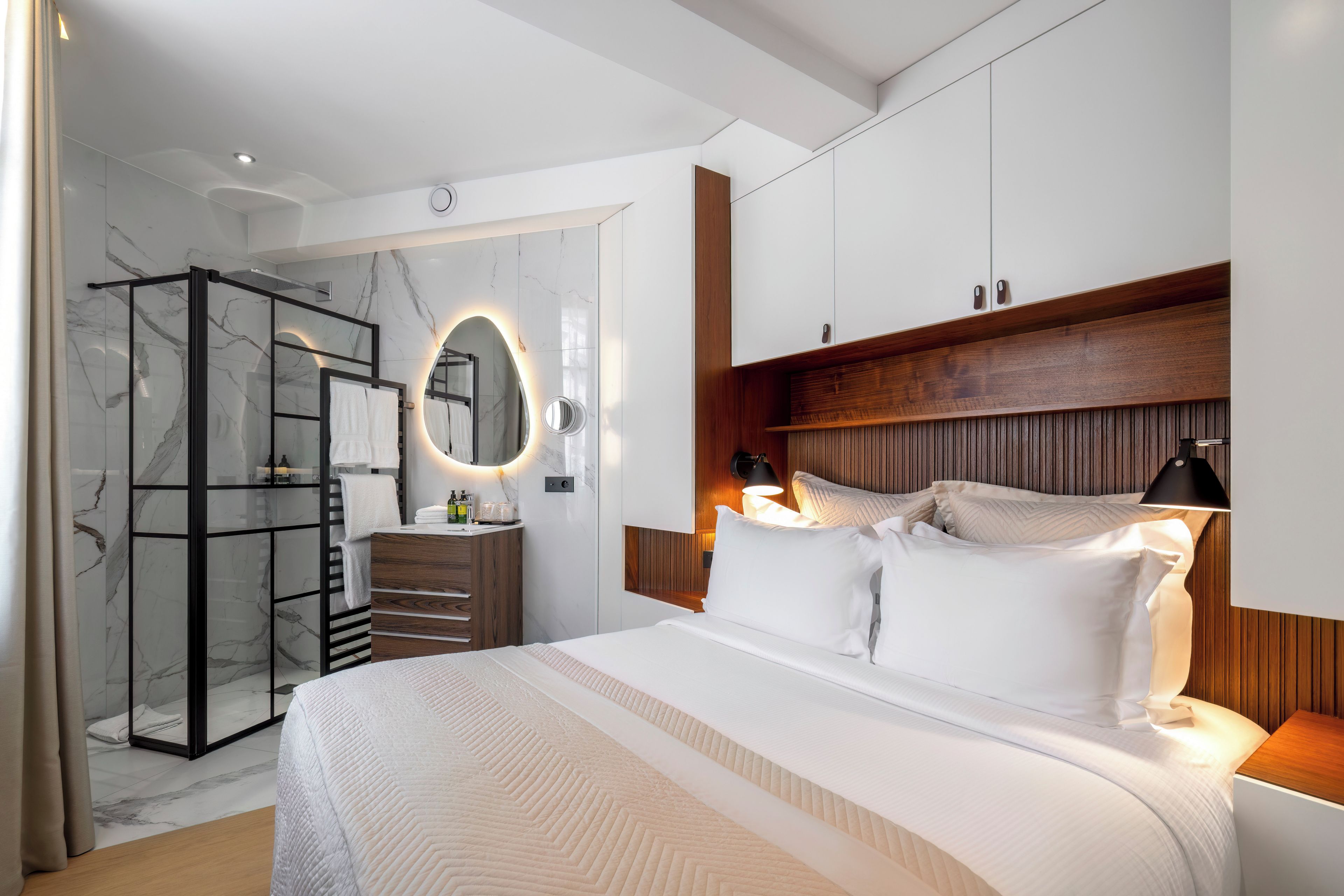 Una camera d'albergo moderna con un letto grande, una doccia a filo pavimento, un angolo trucco con specchio e pannelli in legno sulle pareti.