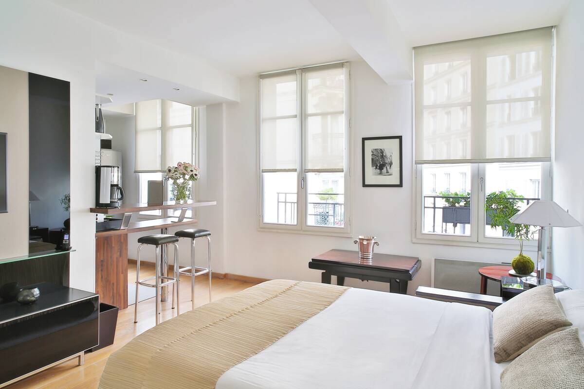 Una habitación de hotel moderna con una cama grande, una kitchenette, un área de descanso con una mesa y sillas, y ventanas amplias con vistas.