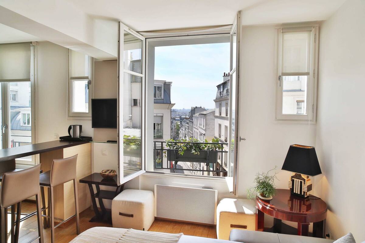 Un apartamento estudio compacto con kitchenette, zona de comedor, sofá, pequeña mesa con lámpara y ventana grande con vistas a la ciudad.