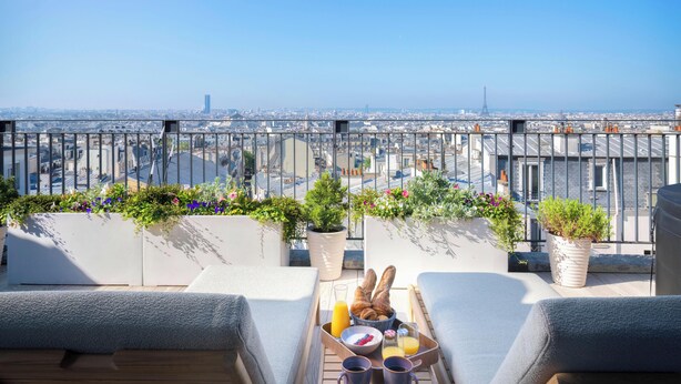 Eine Dachterrasse mit Blick auf die Stadt, Lounge-Sessel, ein Tablett mit Frühstück und Kübelpflanzen.