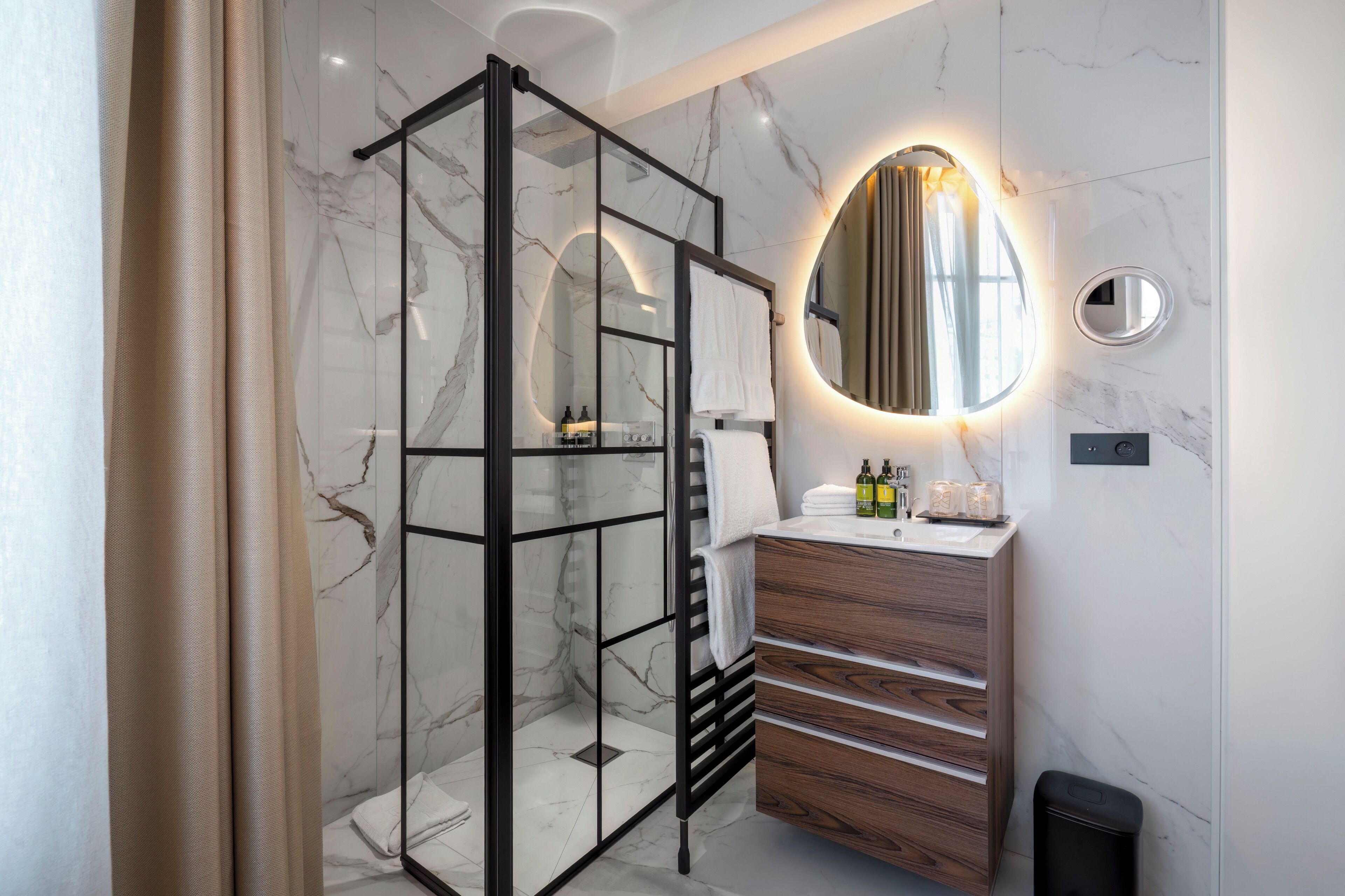 Une salle de bain moderne avec une douche à l’italienne en verre, un meuble-lavabo en bois, un mur recouvert de carreaux de marbre et un miroir rond avec éclairage intégré.
