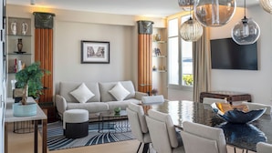 Ein modernes Wohnzimmer mit Sofa, Couchtisch, Esstisch und Stühlen. Es gibt einen Blumentopf, ein gerahmtes Bild an der Wand und einen an der Wand befestigten Fernseher.