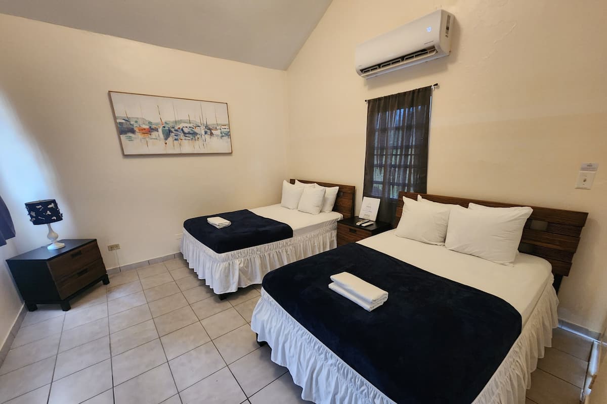 A hotel room with two beds, a painting on the wall, an air conditioner, and a window with curtains.