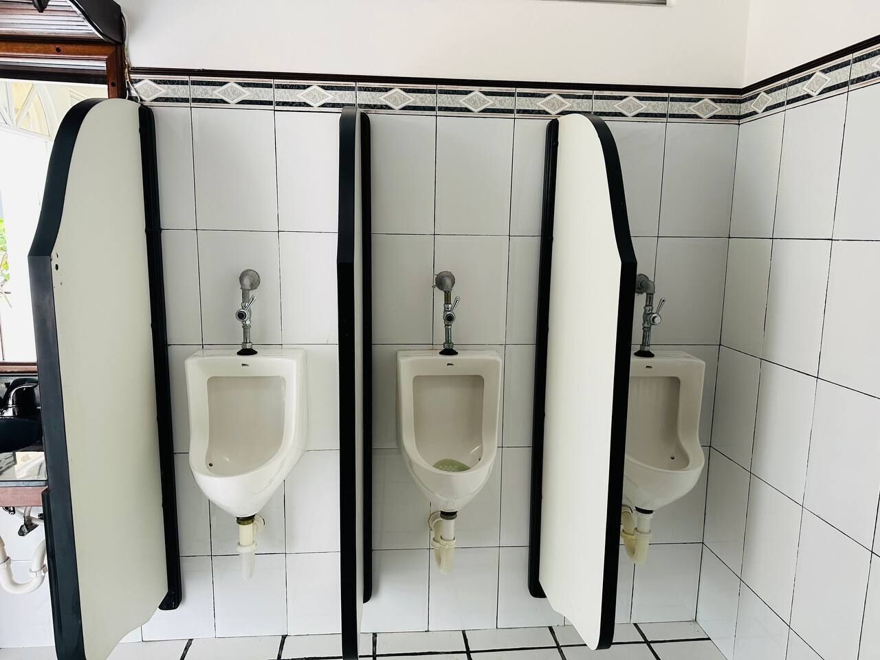 A men's restroom with three urinals, white tiled walls, and black borders.