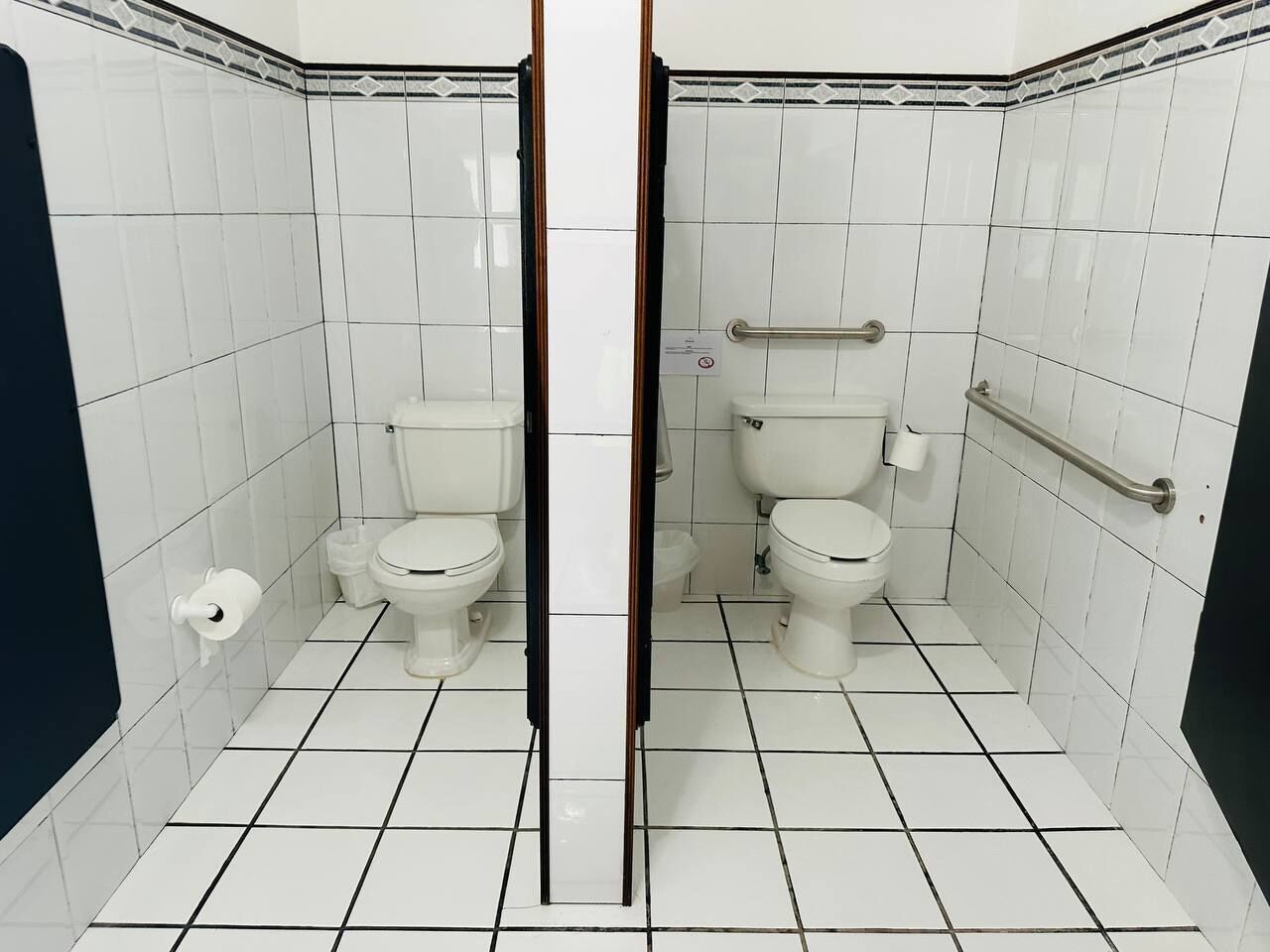 A bathroom with two toilets, white tiled walls, and a handrail.