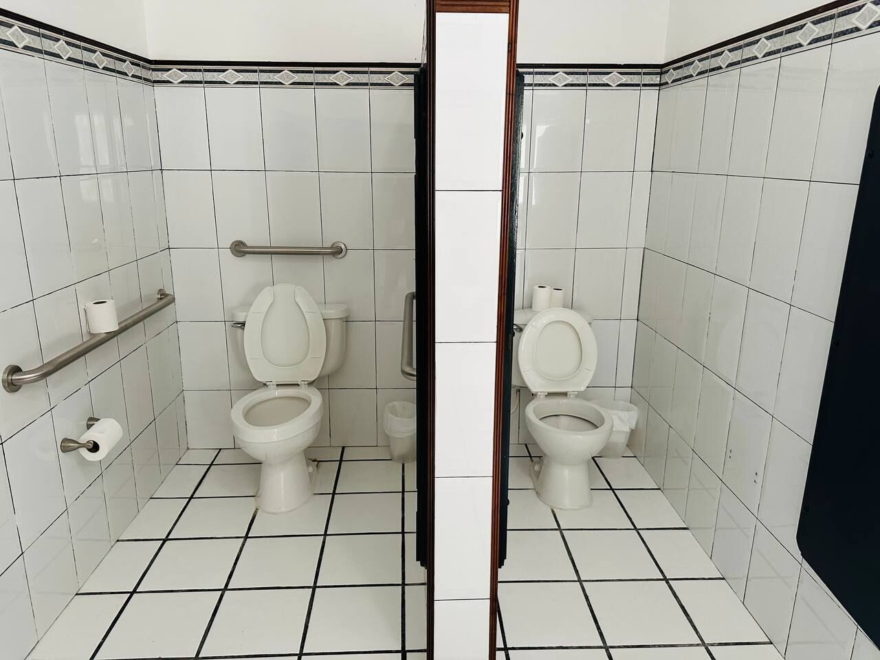 A bathroom with two toilets, a handrail, and a trash bin.