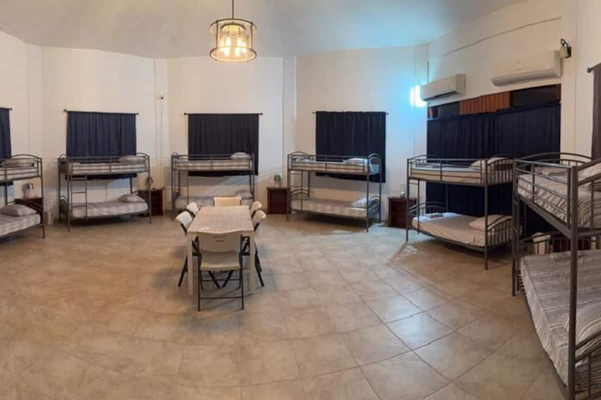 A dormitory room with bunk beds, a table, chairs, a refrigerator, and a fire extinguisher.