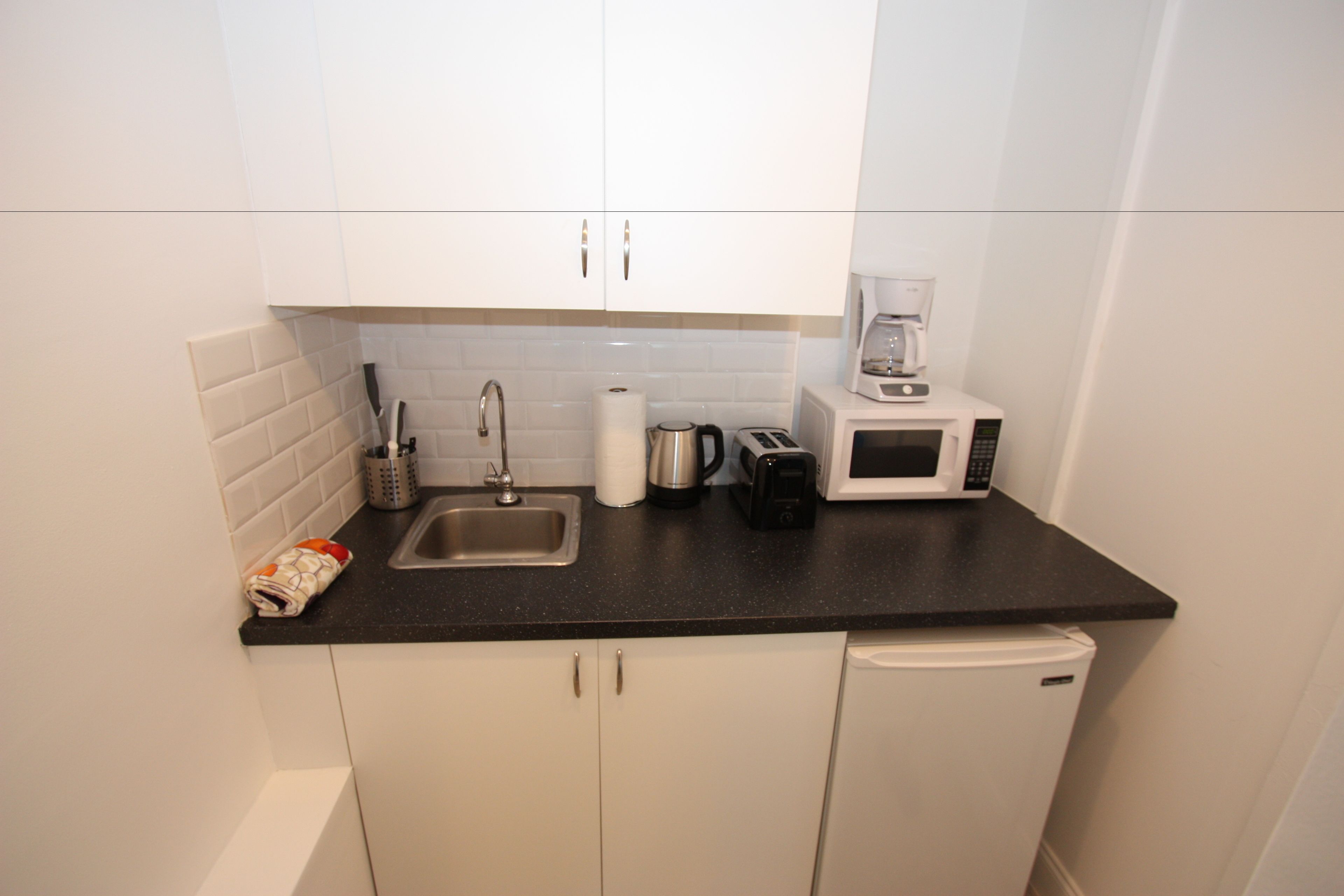 A compact kitchen with white cabinets, a black countertop, a sink, a coffee maker, a toaster, and a small refrigerator.