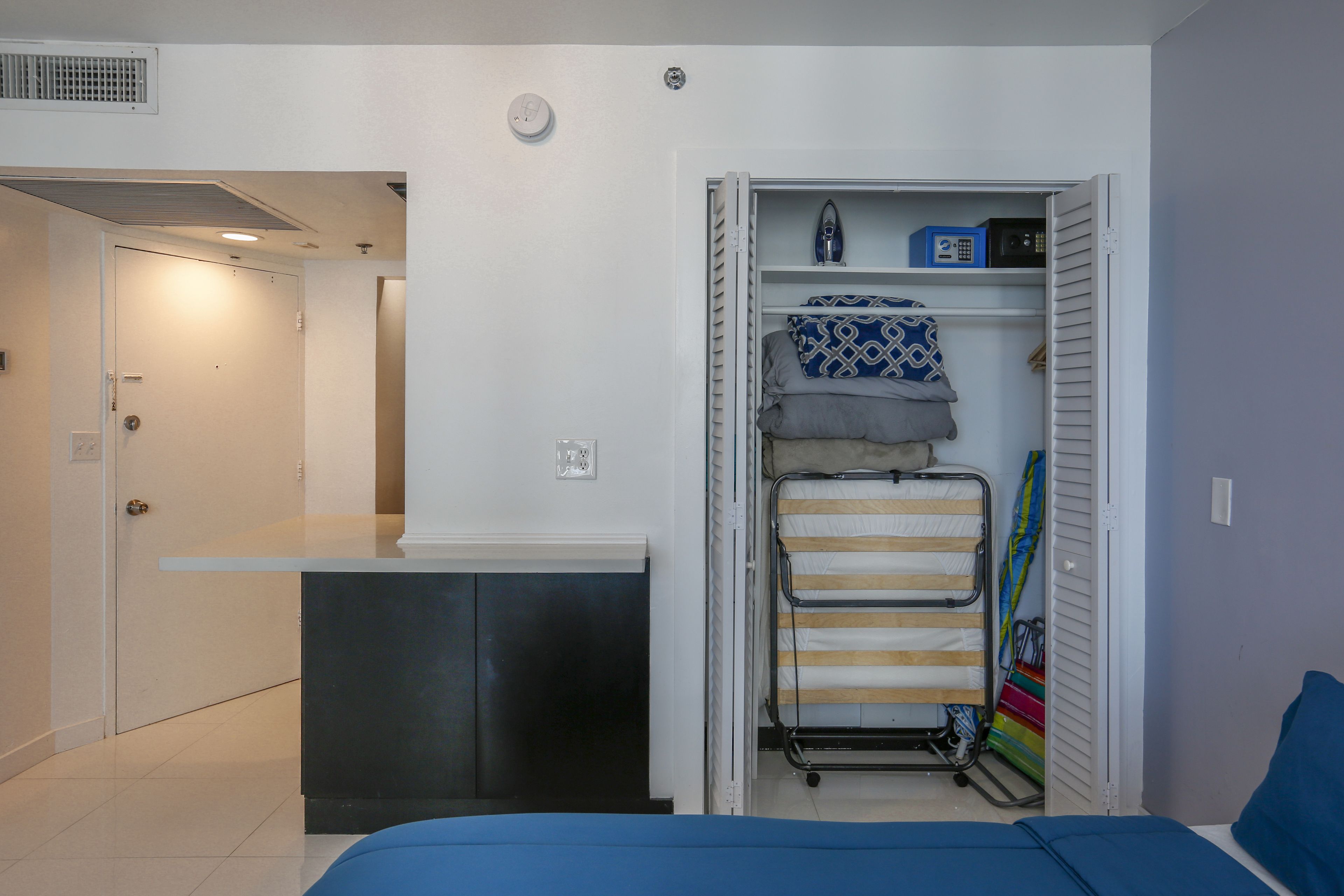 A compact room with a bed, a small desk, a closet with hanging clothes, and a shelf with a radio.