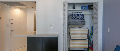 A compact room with a bed, a small desk, a closet with hanging clothes, and a shelf with a radio.