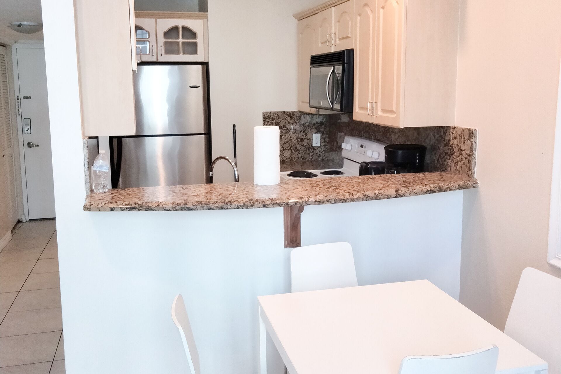 A compact kitchen with a stainless steel refrigerator, a microwave, and a granite countertop. There is a white sink and a small dining table with chairs.