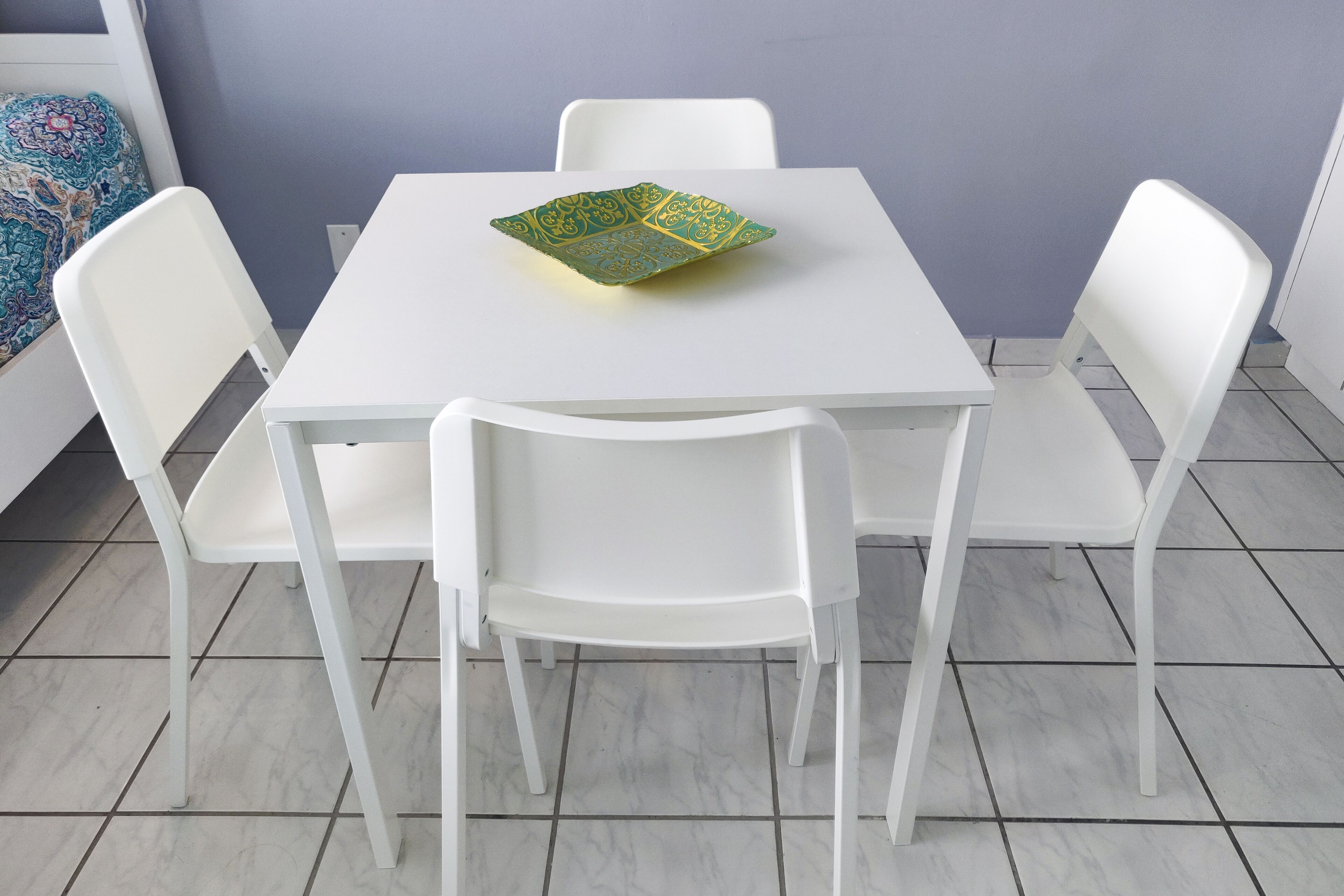 Een witte tafel met een decoratief bord, omringd door witte stoelen, in een kamer met een betegelde vloer en een paarse muur.