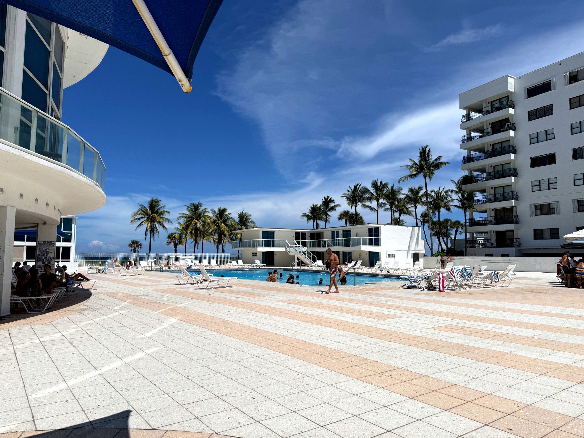 A pool area with lounge chairs, palm trees, and a building with balconies.
