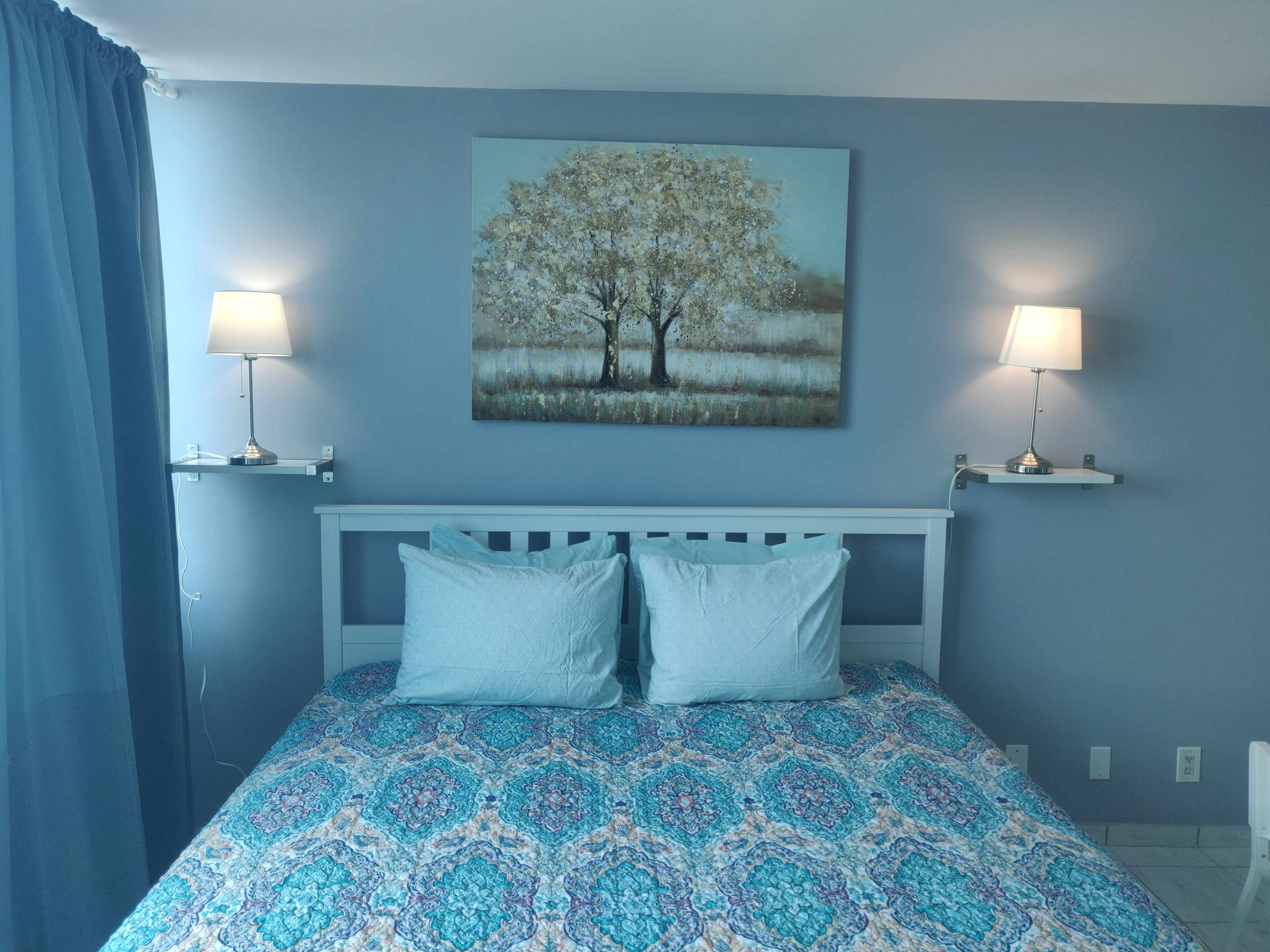 Un dormitorio con una cama, dos lámparas de noche, un cuadro en la pared y una cortina azul.