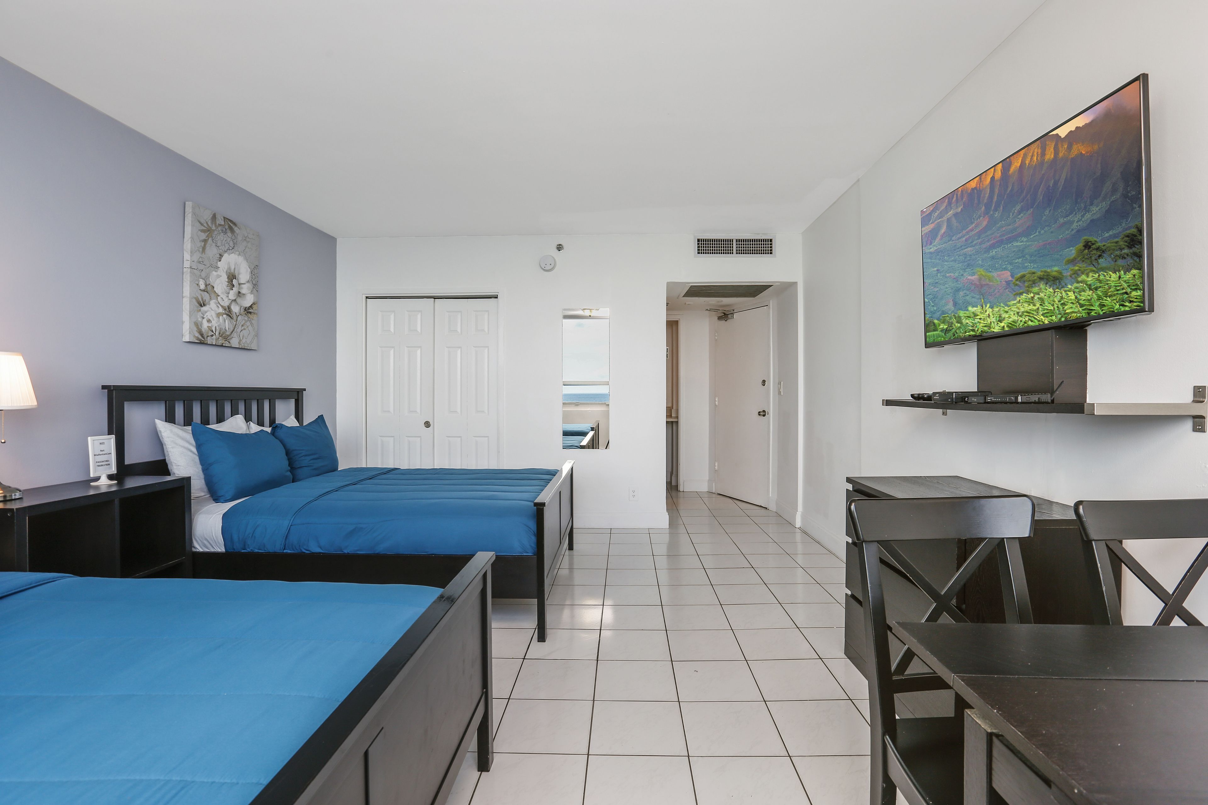 Quarto de hotel com duas camas, uma escrivaninha, uma cadeira, televisão de tela plana e um quadro na parede.