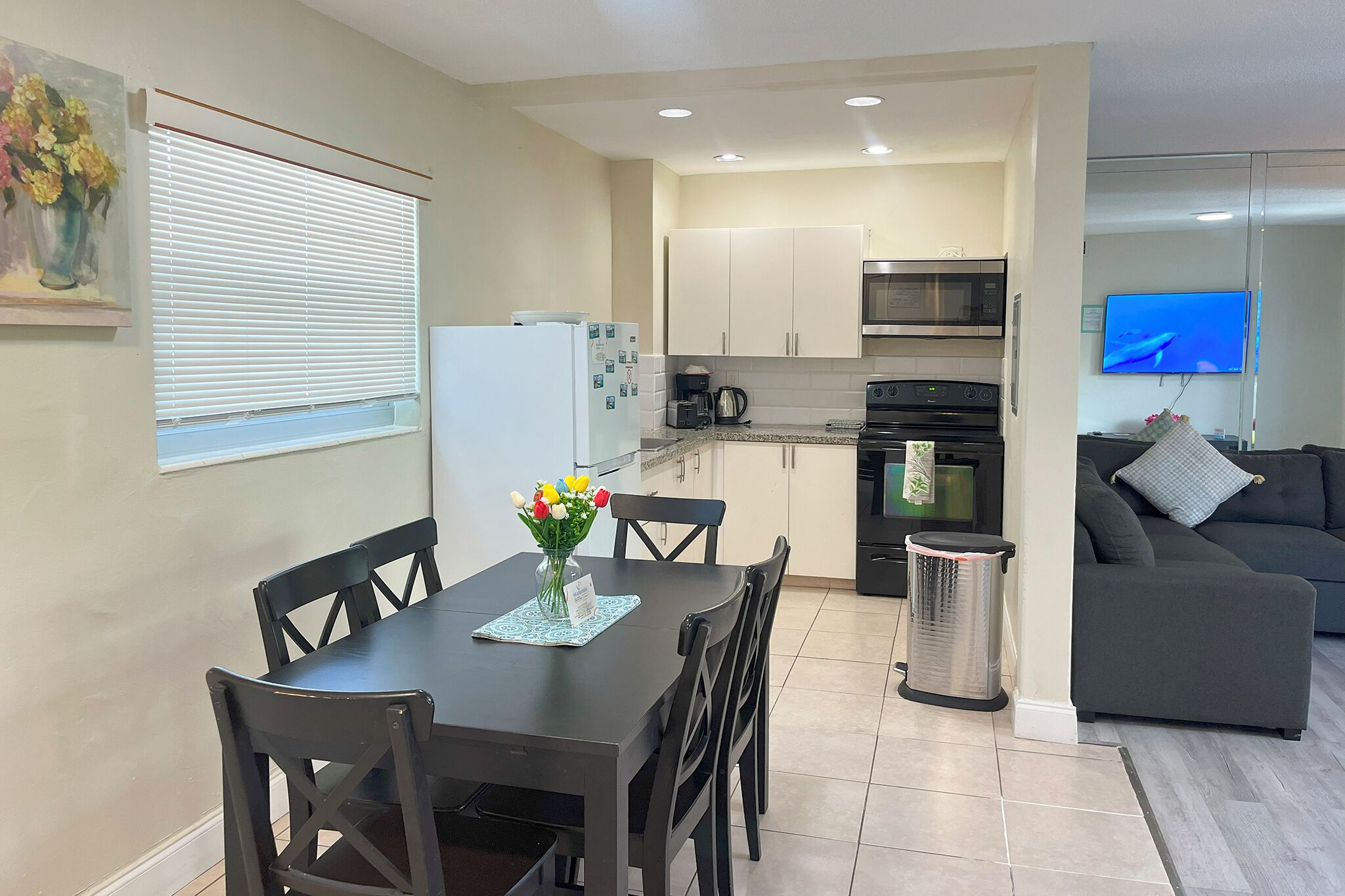 A modern kitchen with white cabinets, a microwave, and a coffee maker. There is a dining table with chairs and a vase of flowers. A black couch and a TV are visible in the living area.