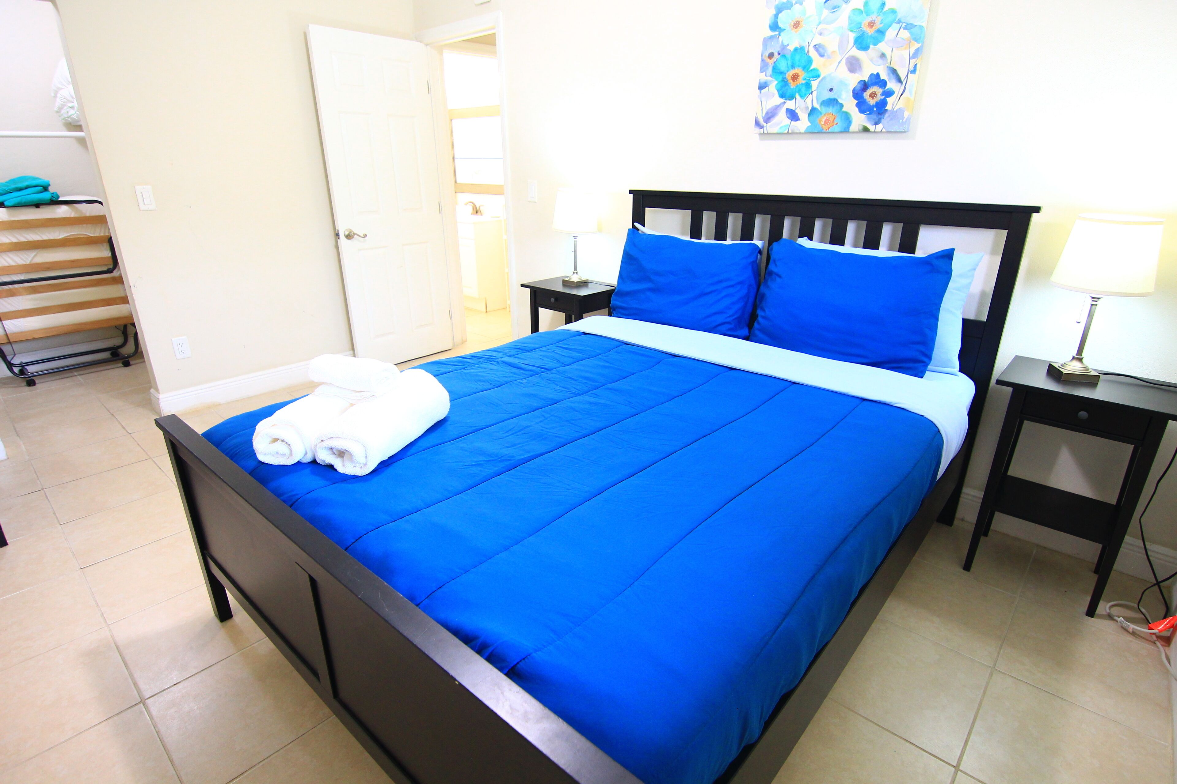 A neatly made bed with blue bedding, a bedside table with a lamp, and a painting on the wall.